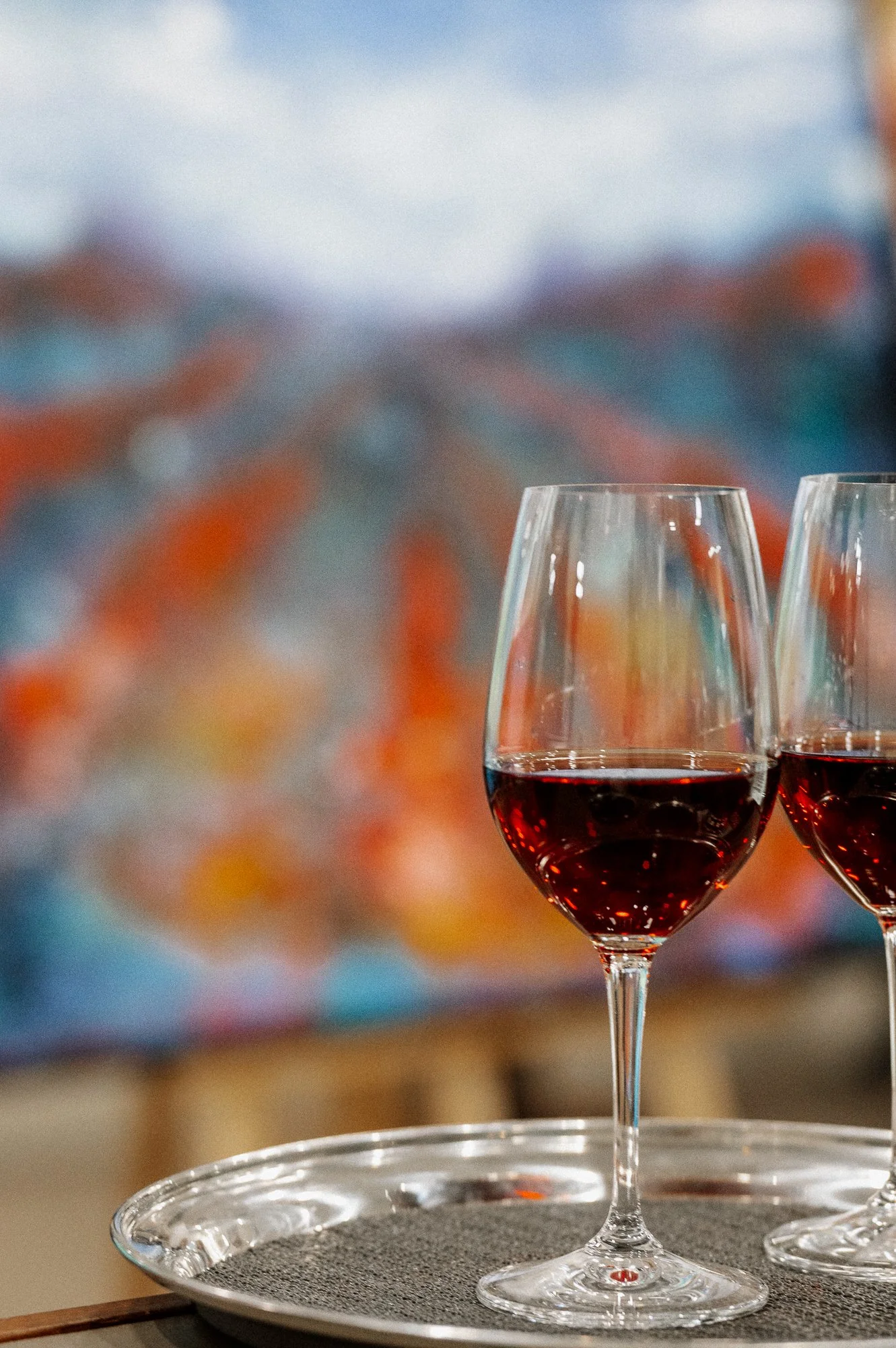 Two glasses of red wine on a silver tray with a blurred colorful background.