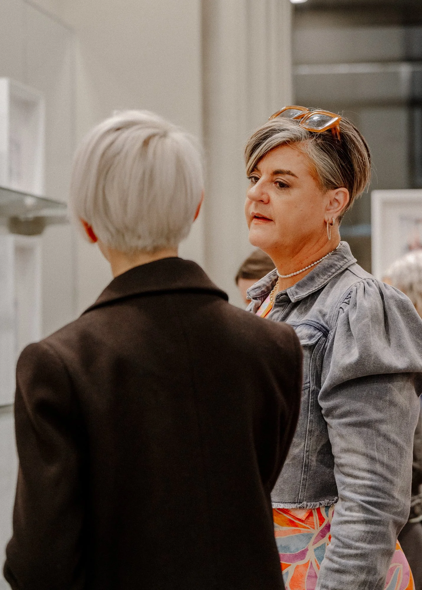 Two women having a conversation indoors. One woman seen from behind with short gray hair, wearing a dark jacket. The other woman facing her with a short hairstyle, glasses on her head, and wearing a denim jacket and colorful dress.