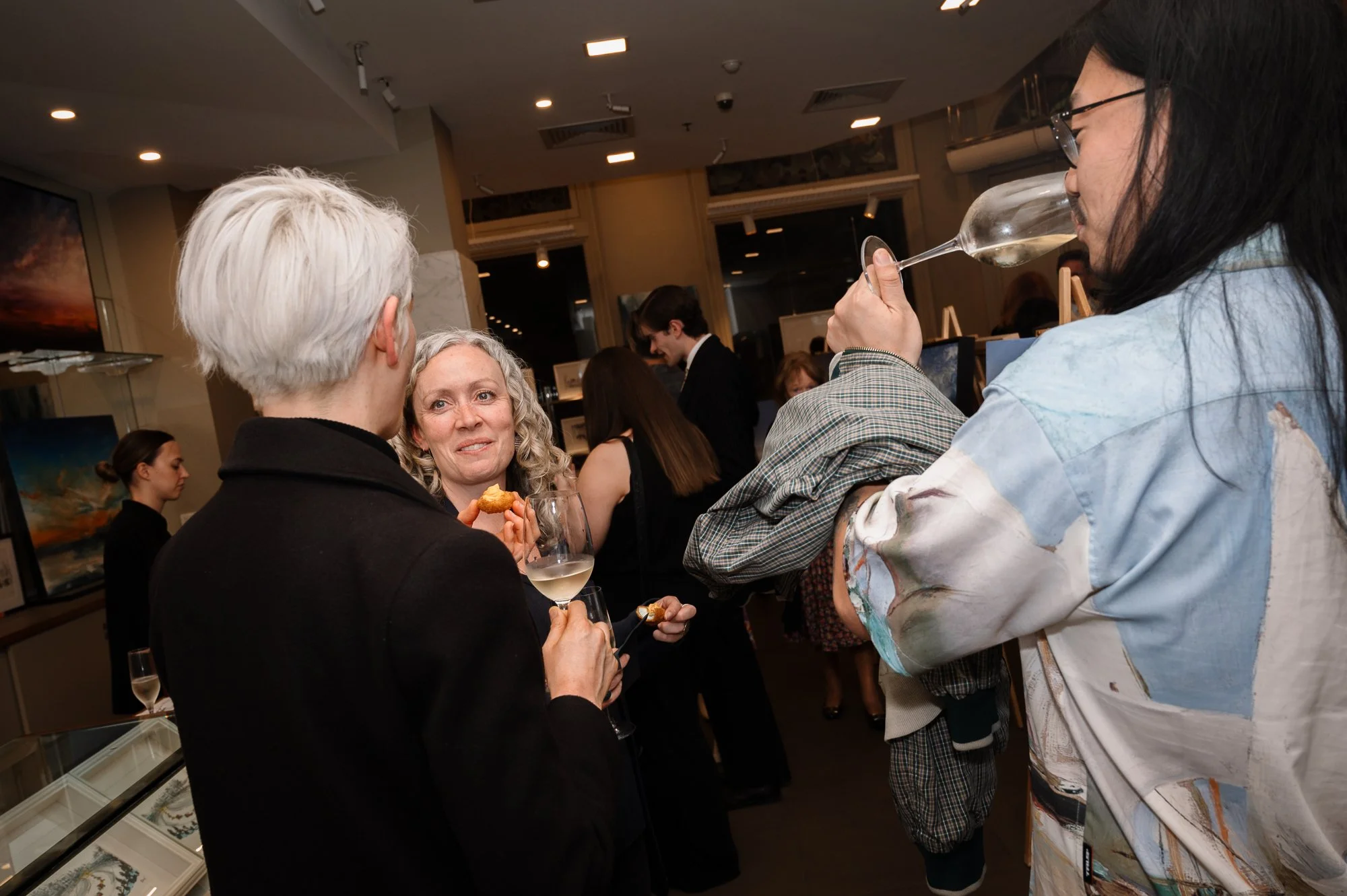 People socializing at an art gallery or exhibition, with some holding glasses of wine and snacks.