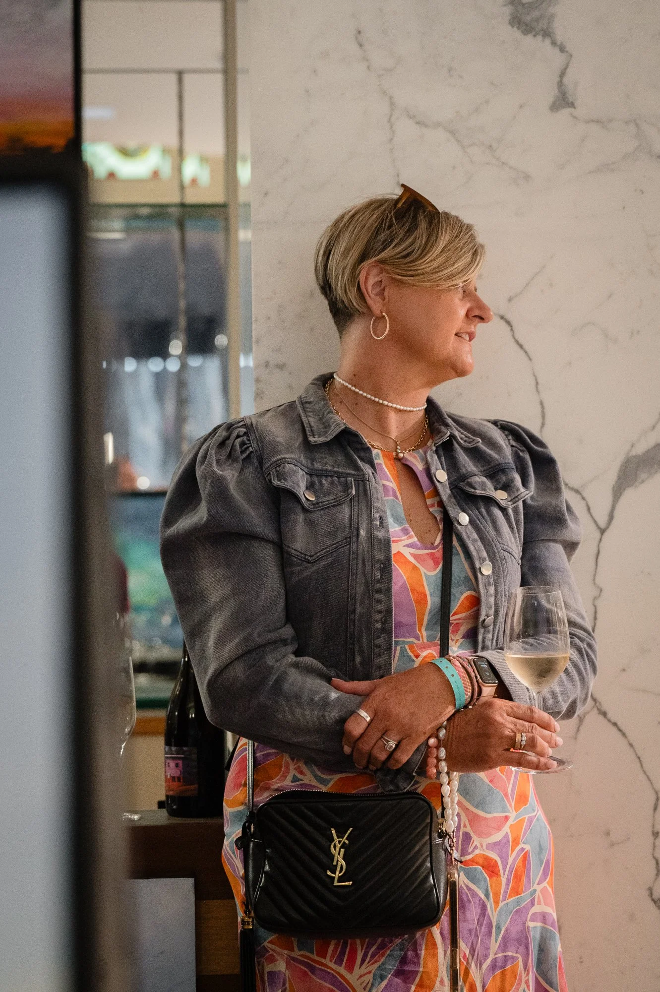 A middle-aged woman with short blonde hair, wearing earrings, a necklace, and a pearl bracelet, holds a glass of white wine. She is dressed in a colorful patterned dress and a gray denim jacket. She has a black Yves Saint Laurent purse hanging from h