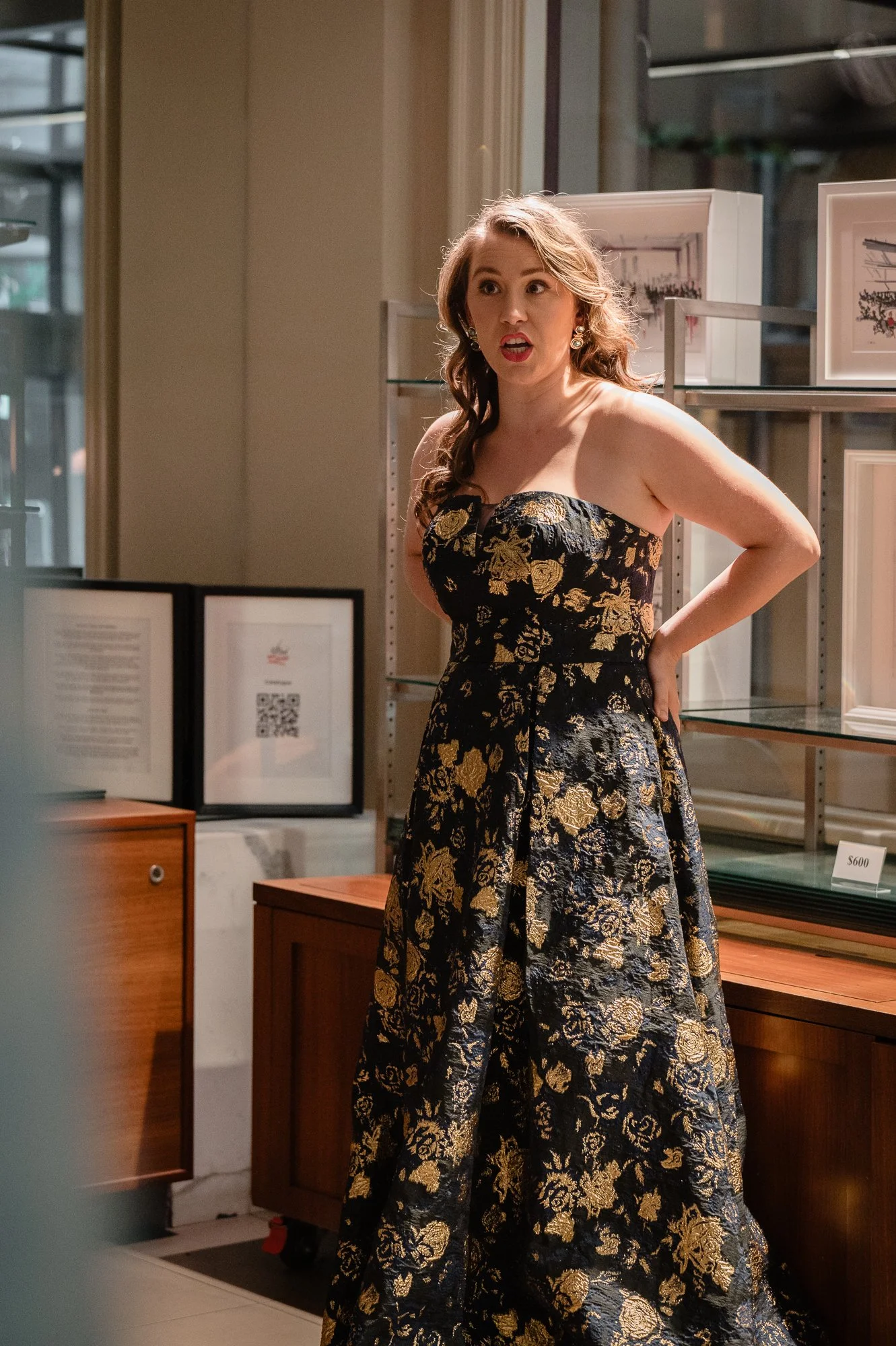 A woman in a black and gold floral strapless dress stands with her hand on her hip, making an expressive face, inside an art gallery or store with framed pictures on shelves behind her.