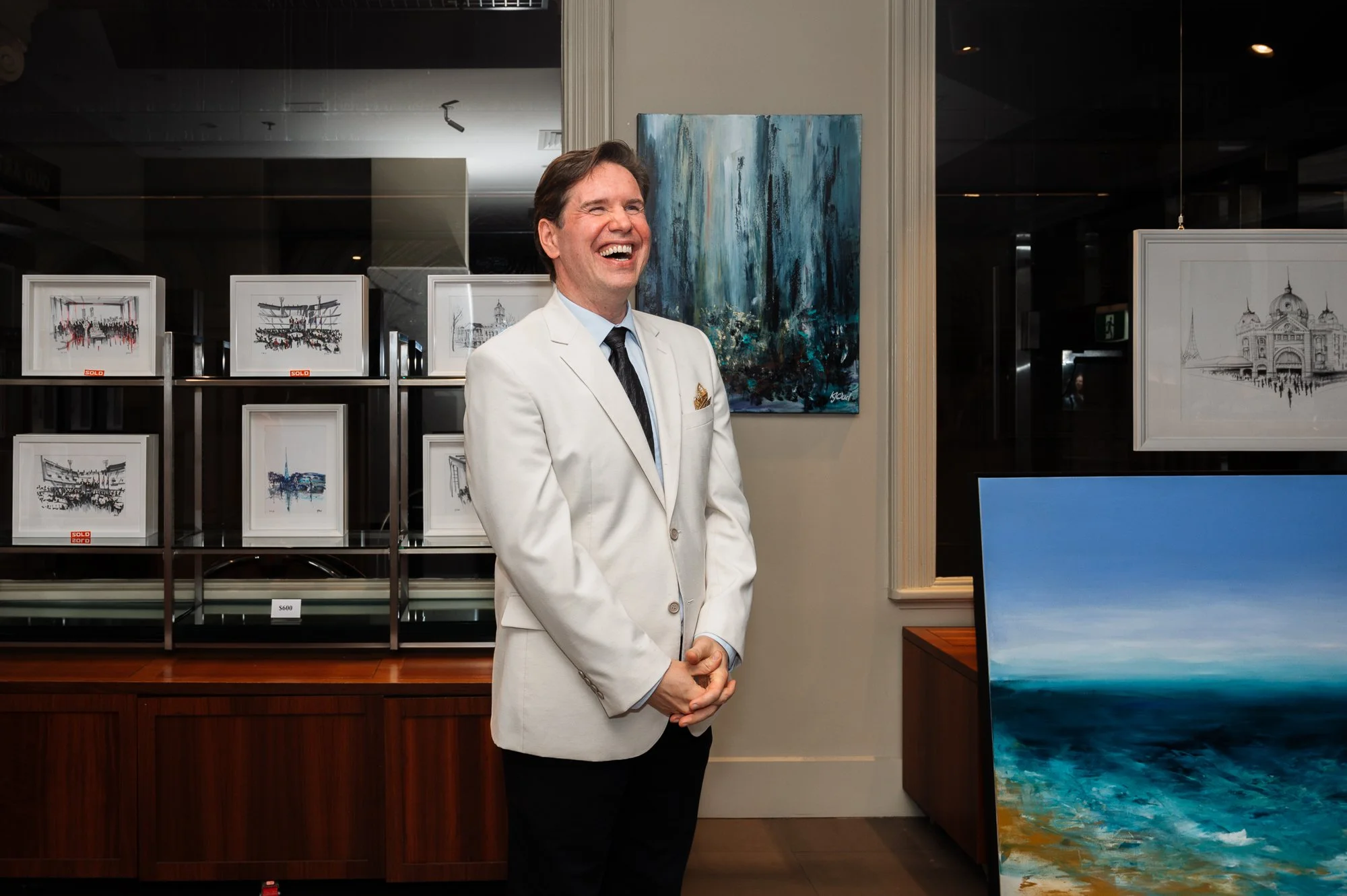 A man in a white suit jacket and black tie standing and smiling in an art gallery with several framed artwork behind him and a large abstract painting to his right.
