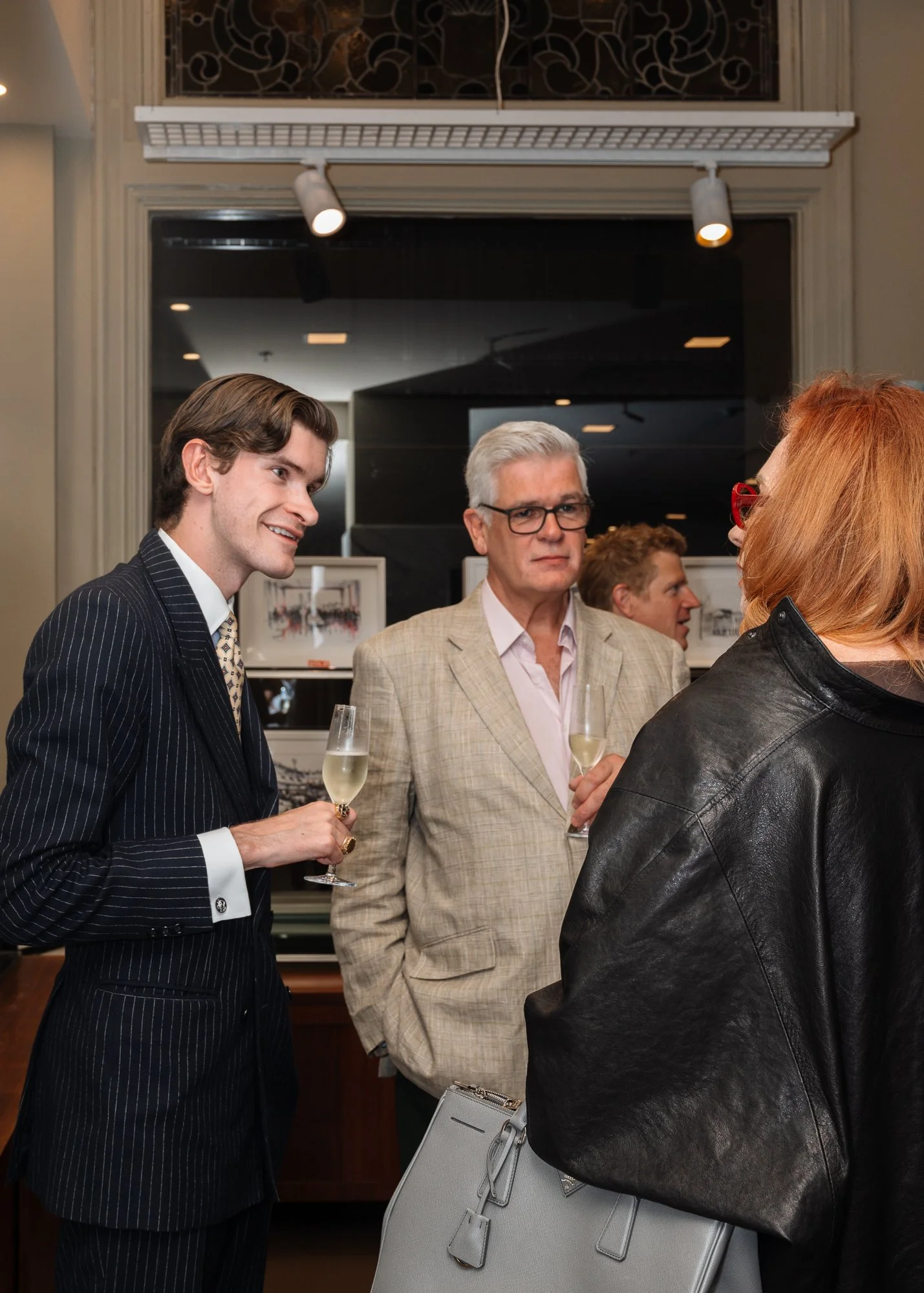 People at a social event are holding glasses of champagne and engaging in conversation. A woman with red hair and black leather jacket is facing away, while young and older men are facing her, dressed in formal and semi-formal attire.