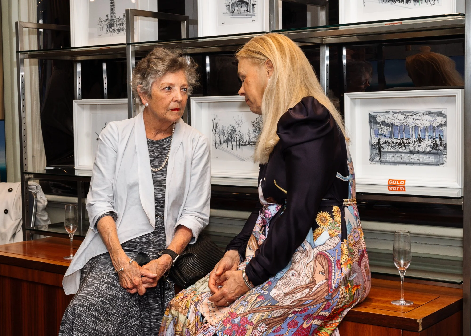 Two women having a conversation in an art gallery, with framed artwork behind them and champagne glasses on a table.