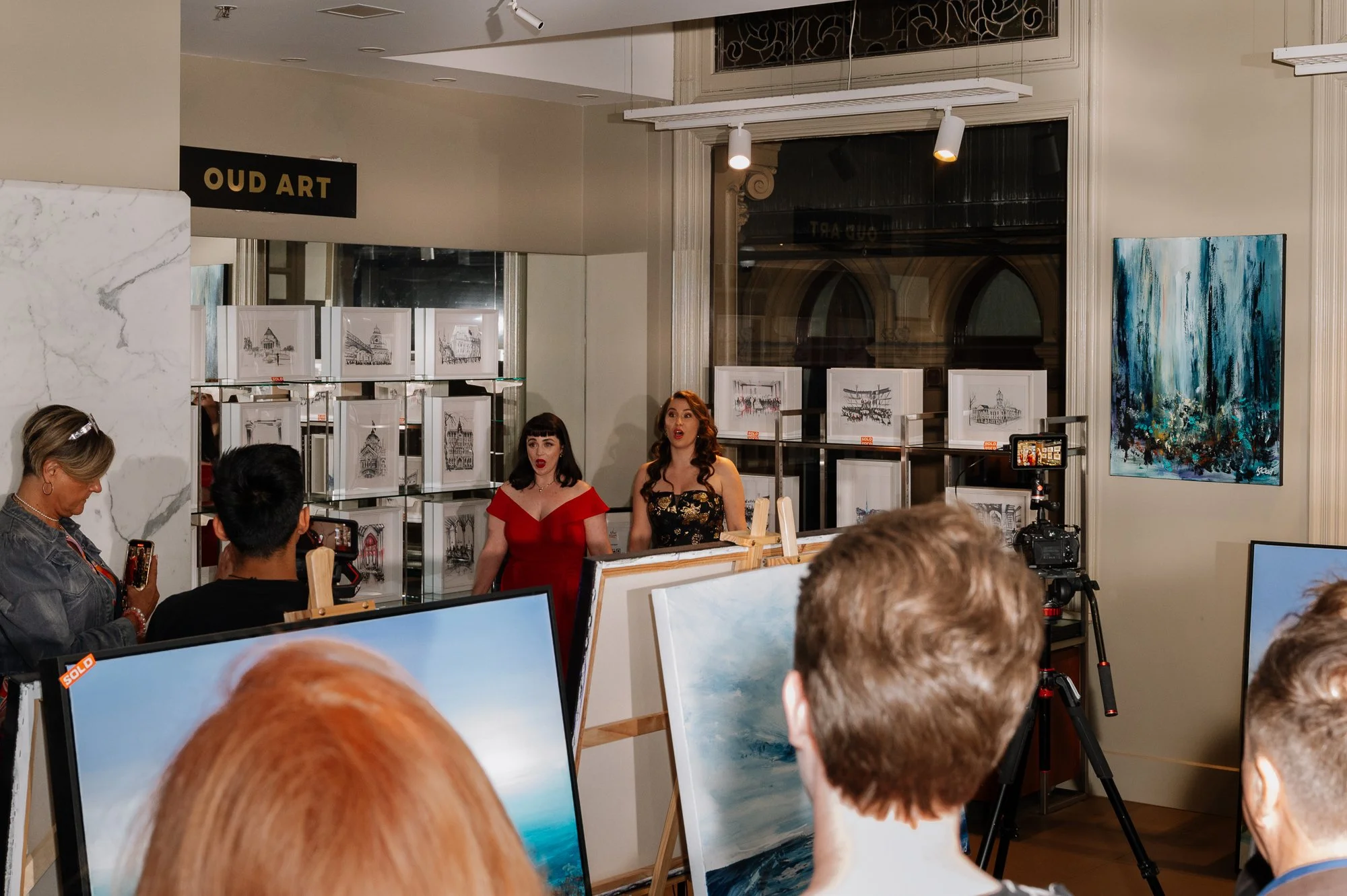 Two women singing or speaking at an art gallery event with artwork displayed behind them and an audience watching.