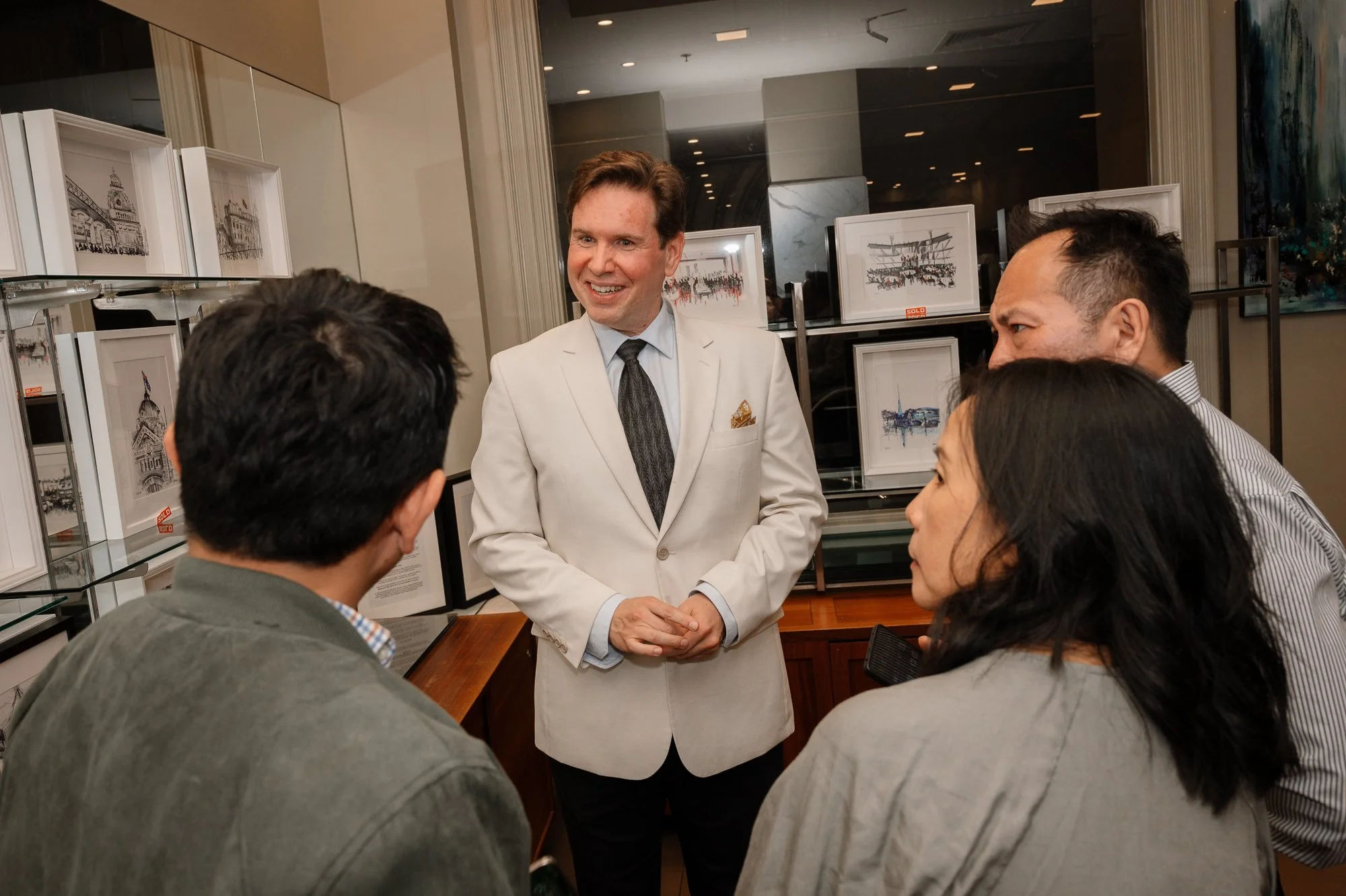 A man in a white blazer speaking to a group of four people in an art gallery or exhibit space, with framed artwork displayed behind them.
