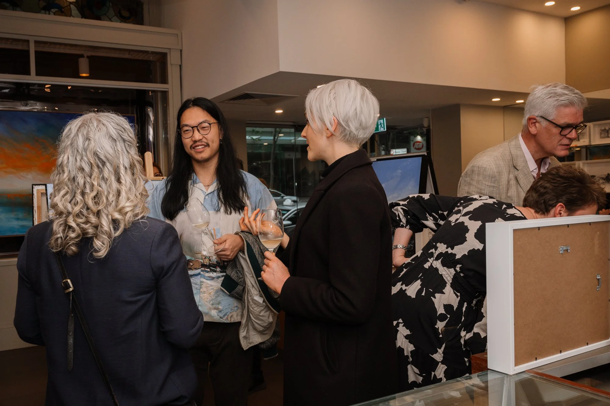 Group of people socializing at an art gallery or exhibition, with paintings displayed in the background, some holding wine glasses.