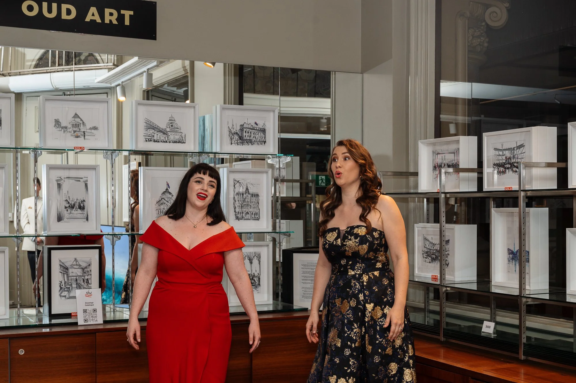 Two women in formal dresses standing inside an art gallery or exhibit with framed sketches of buildings displayed on glass and metal shelves behind them.