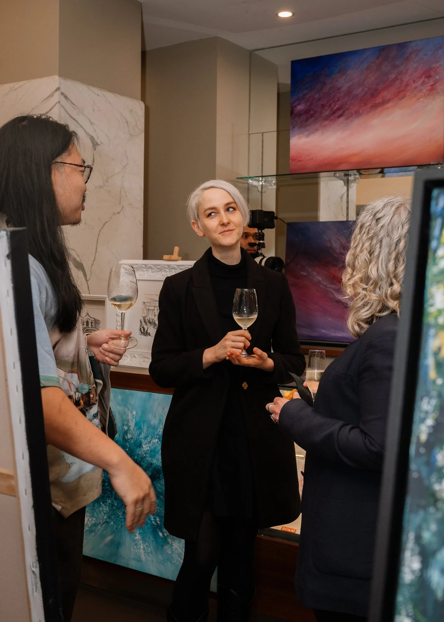 Three women are engaged in conversation at an art gallery, holding glasses of white wine. Background features abstract paintings and art displays.