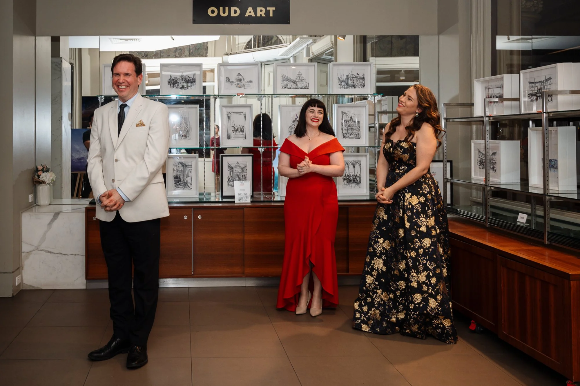 Three people standing inside an art gallery with framed black-and-white artwork displayed behind them. The man on the left is wearing a white tuxedo jacket, black pants, and black shoes, and is smiling. The woman in the middle is wearing a red off-th