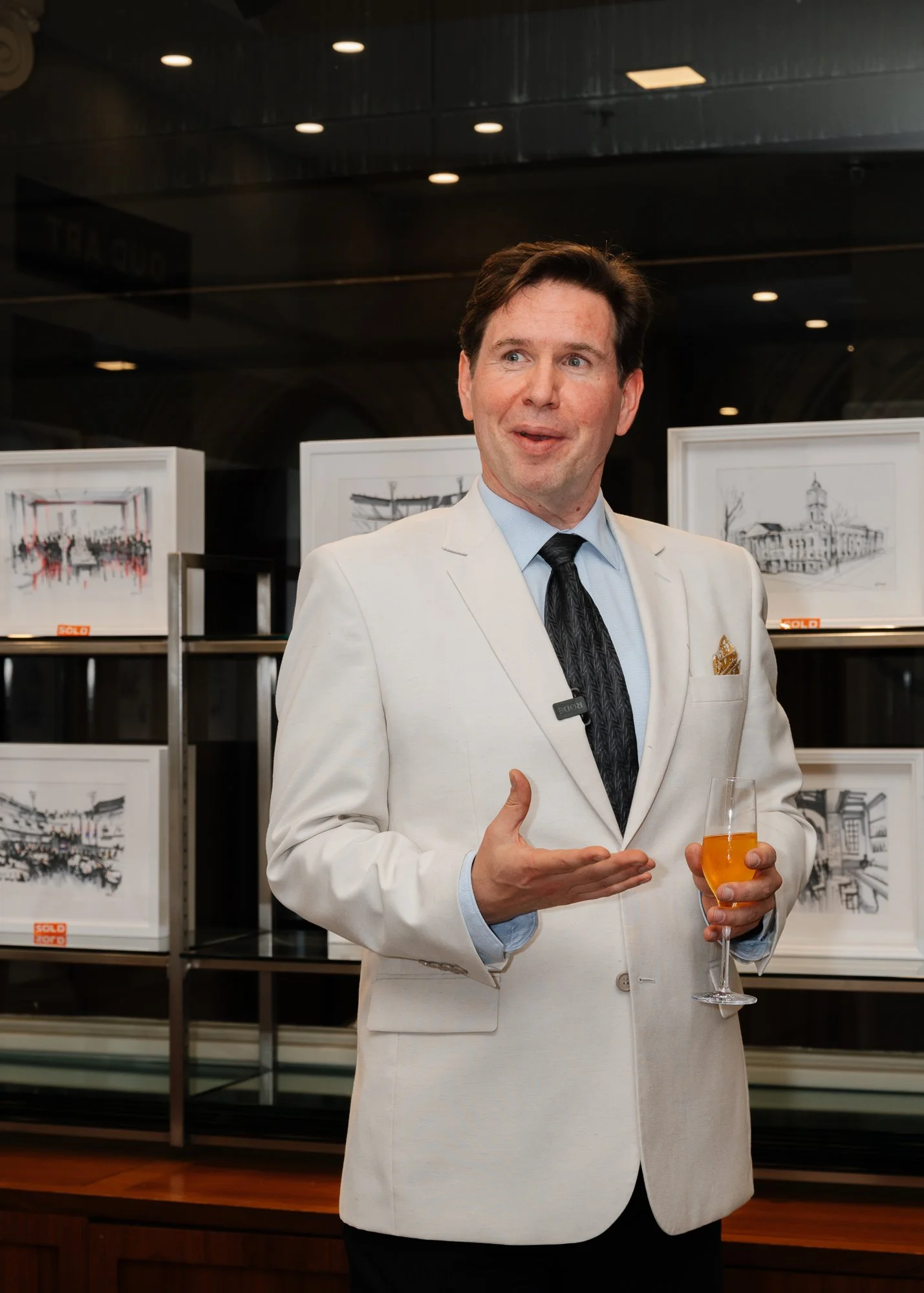 Man in white suit speaking, holding a glass of drink, behind framed artwork at an art gallery.