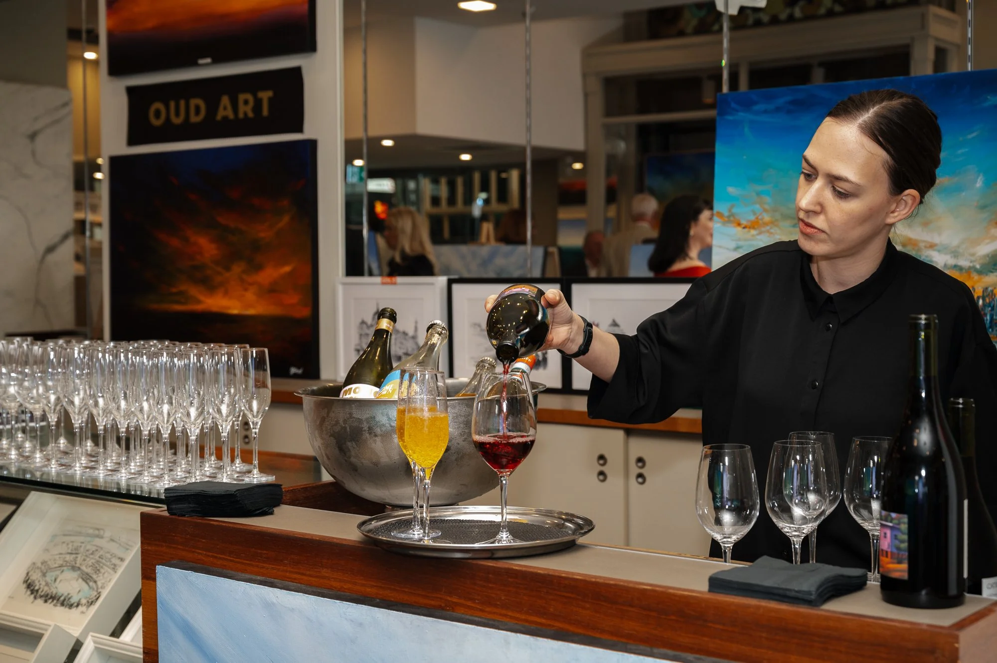 A bartender pouring red wine into a glass at a bar area with art paintings on the wall behind her and a tray of glasses on the counter.