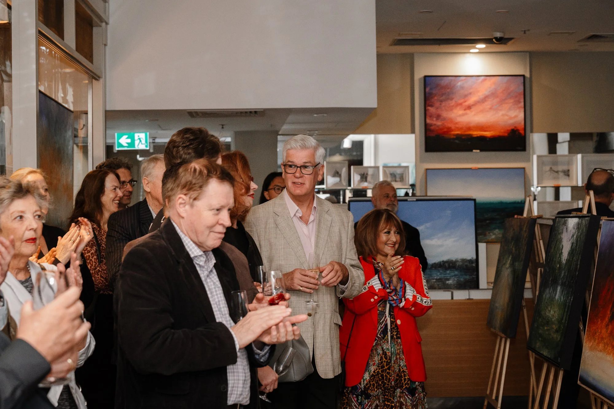 A group of people at an art gallery, observing and applauding paintings on display, holding drinks, with some smiling and engaging in conversation.