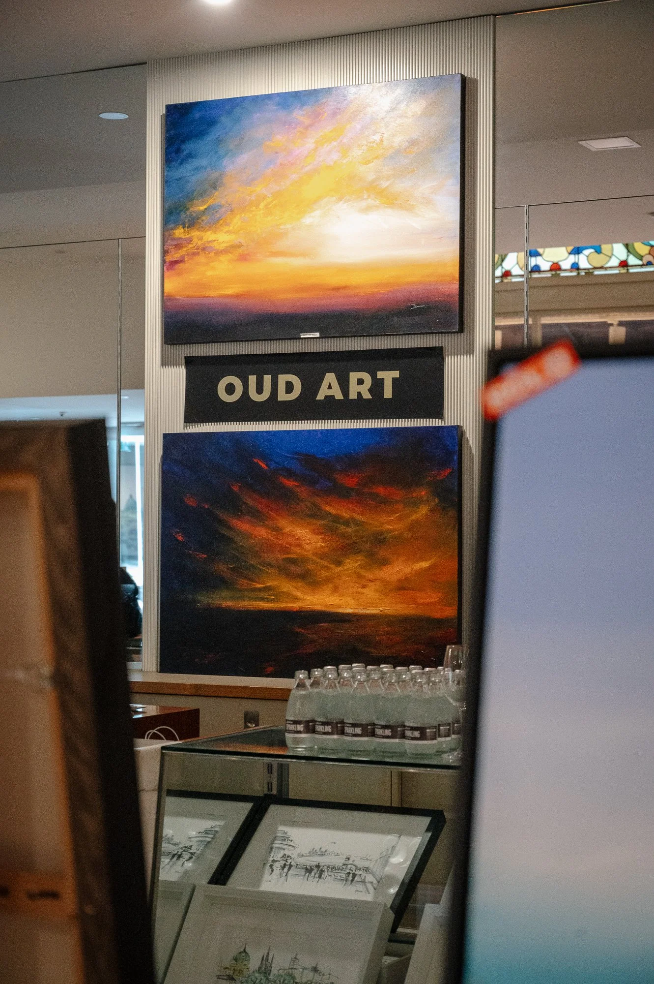 Two landscape paintings of sunset skies displayed on a wall, with a sign that reads 'OUD ART' between them.