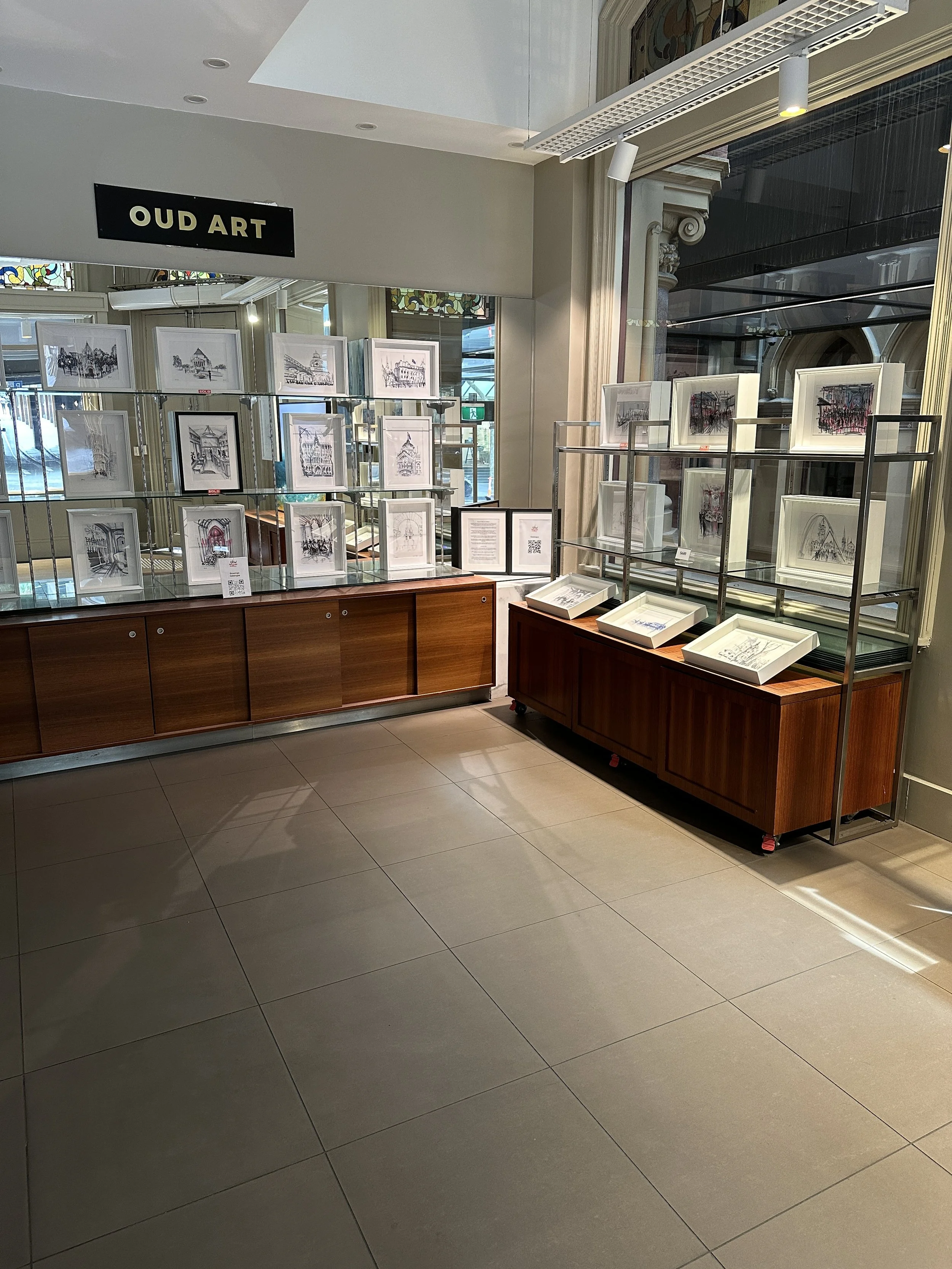 Art exhibit titled 'OUD ART' with framed drawings or paintings displayed on glass and metal shelves inside a gallery with large windows and tiled floor.