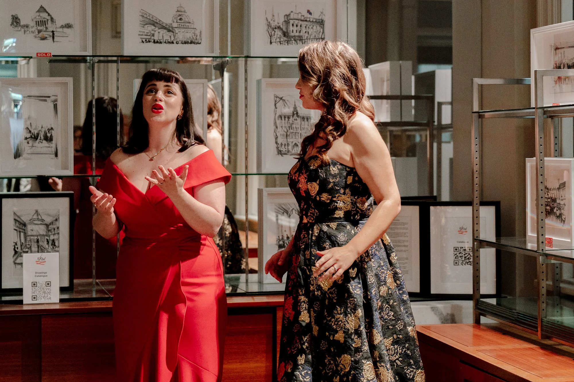 Two women are talking at an art gallery, one wearing a red dress with off-the-shoulder sleeves, and the other wearing a black dress with gold floral patterns. There are framed drawings of cityscapes or architecture displayed on glass shelves behind t