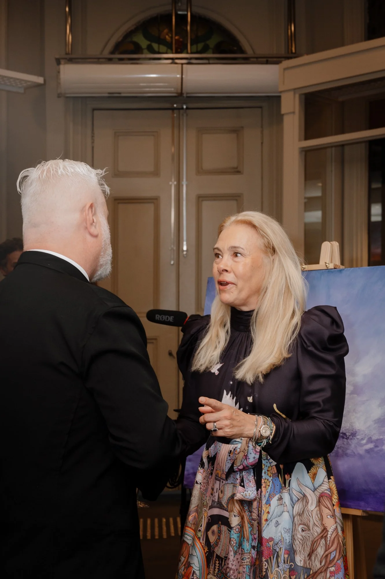 A woman with long blonde hair and a colorful illustrated skirt is talking to a man with short white hair, dressed in a black suit. They are indoors near an art piece and a microphone.