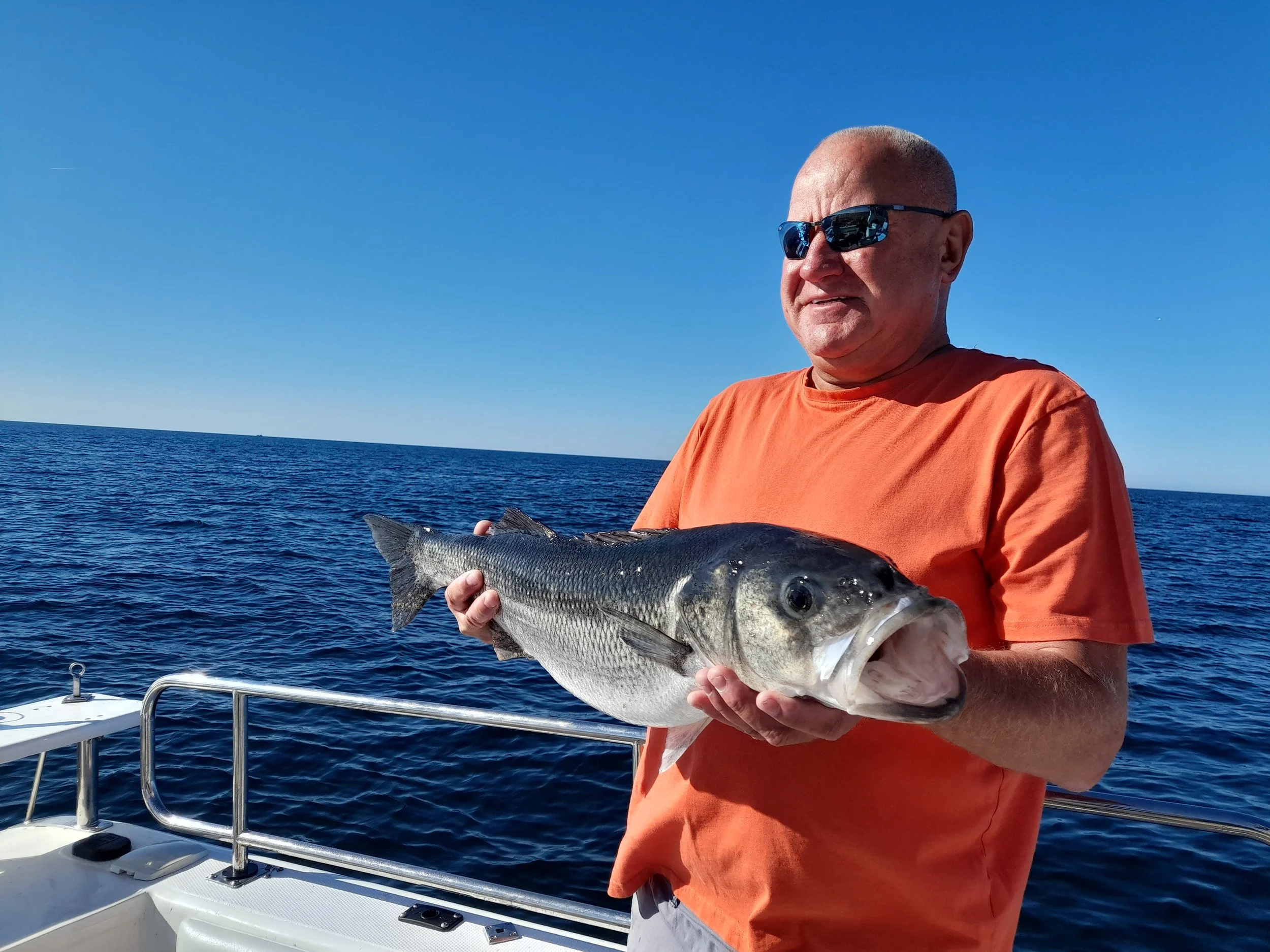 Man in orange shirt and sunglasses holding a large fish on a boat at sea with a clear blue sky.
