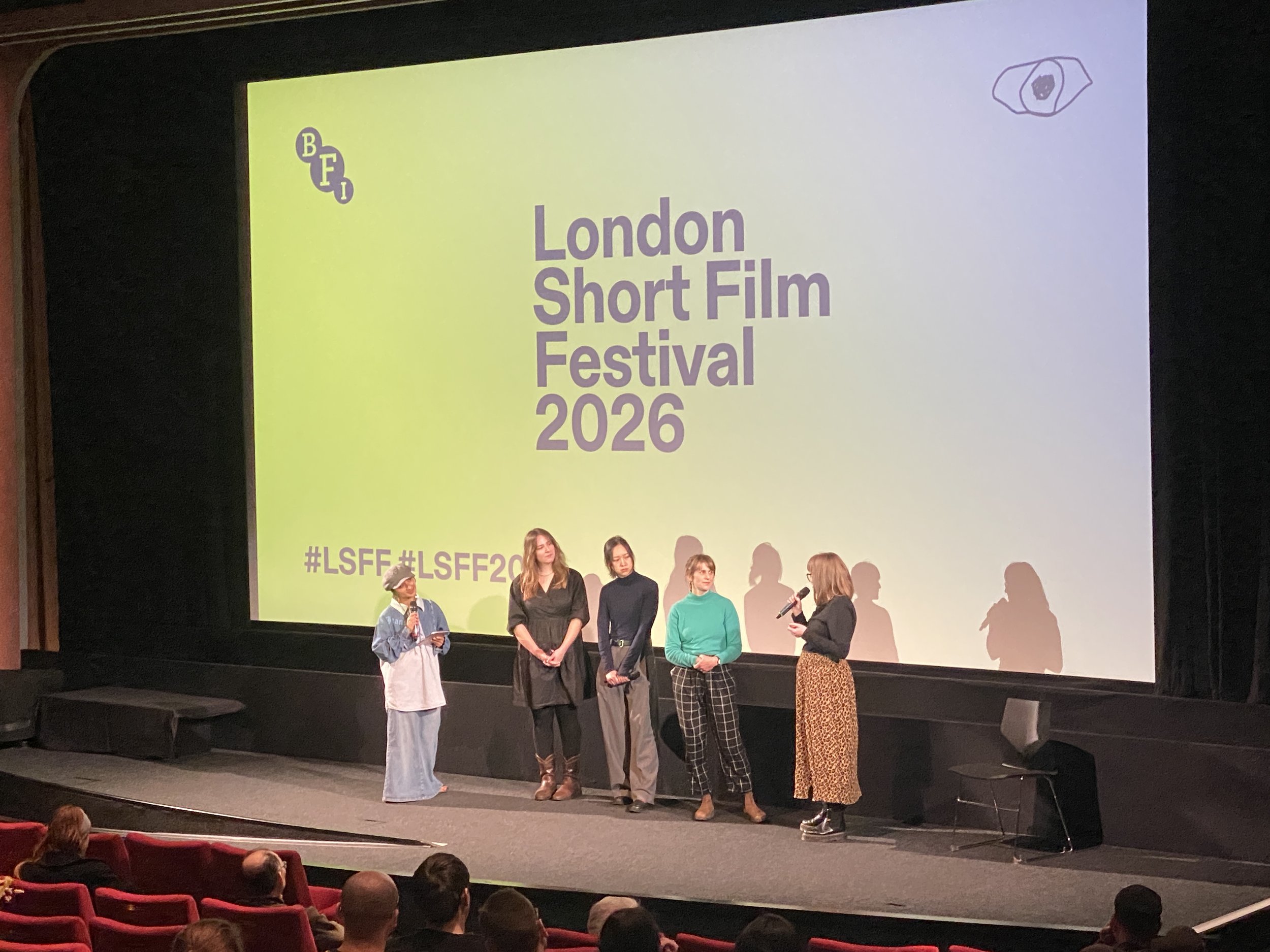 Esther is standing on a stage with a microphone and a group of people. The cinema screen behind her says "London Short Film Festival 2026"