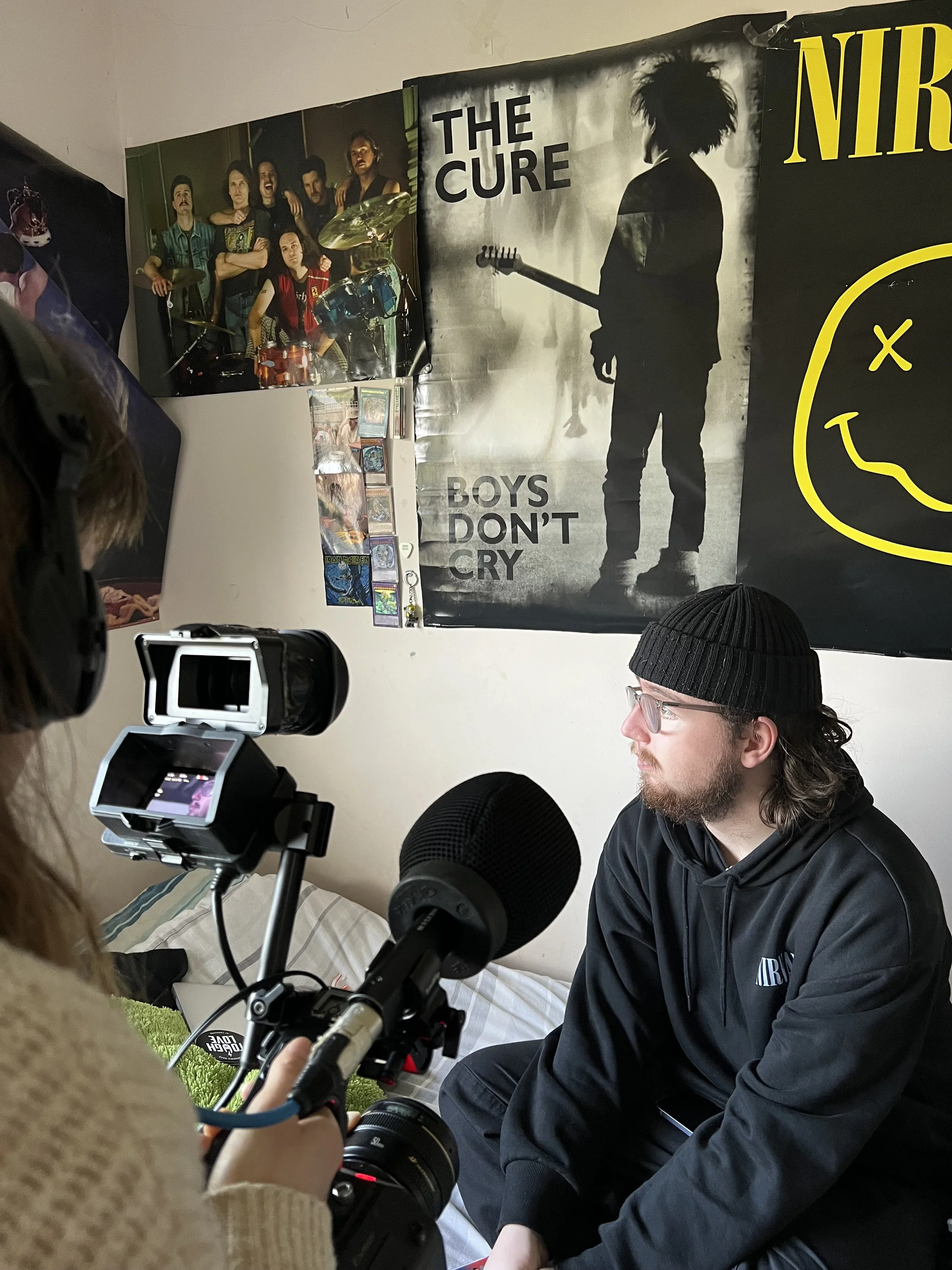 A man wearing a black beanie hat and hoodie is sitting on a bed with posters on the wall behind. Someone holding a microphone stands partly off camera.