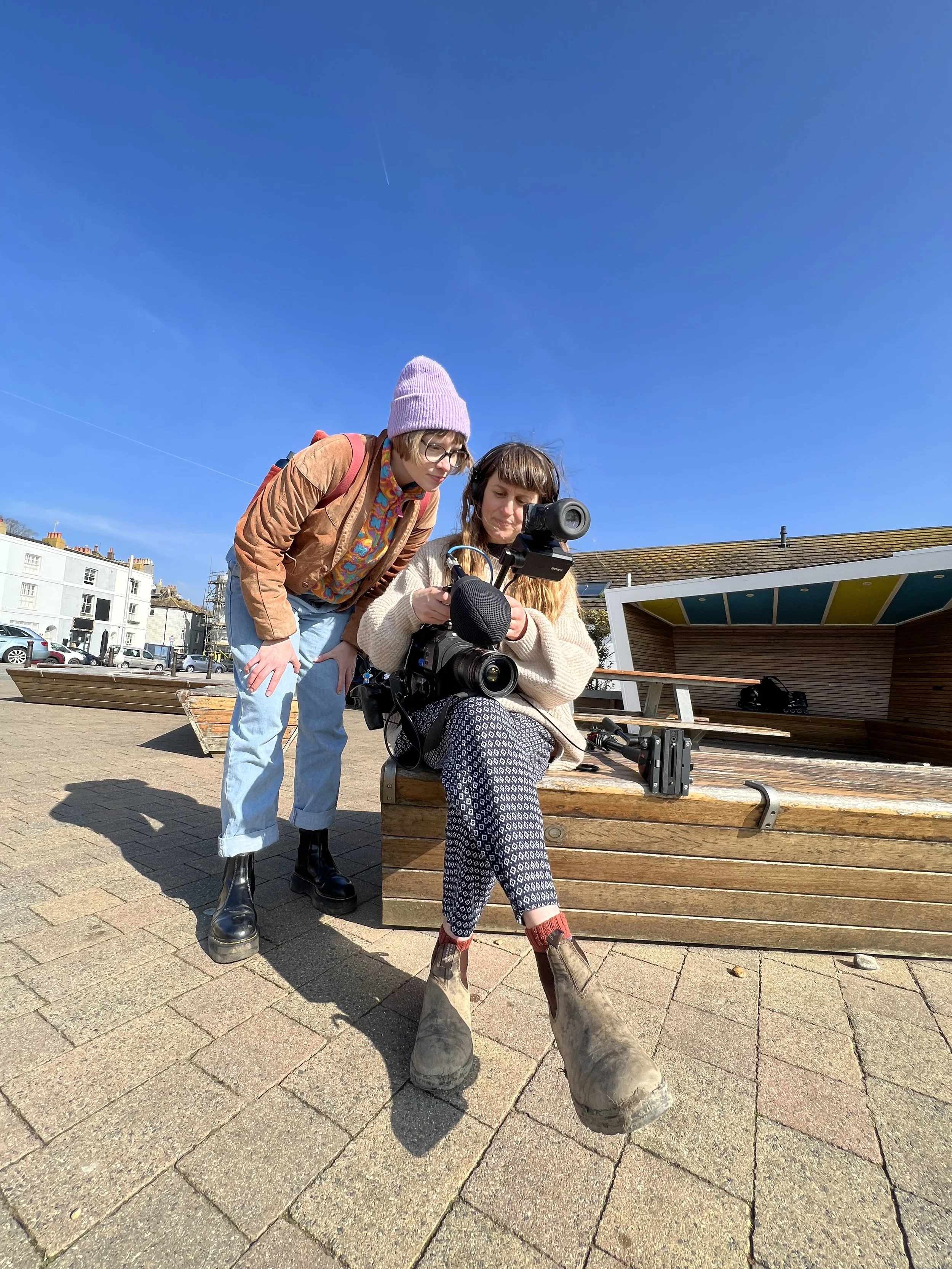 Esther is standing behind a young person holding a camera and looking over her should to see what is being shown in the viewer