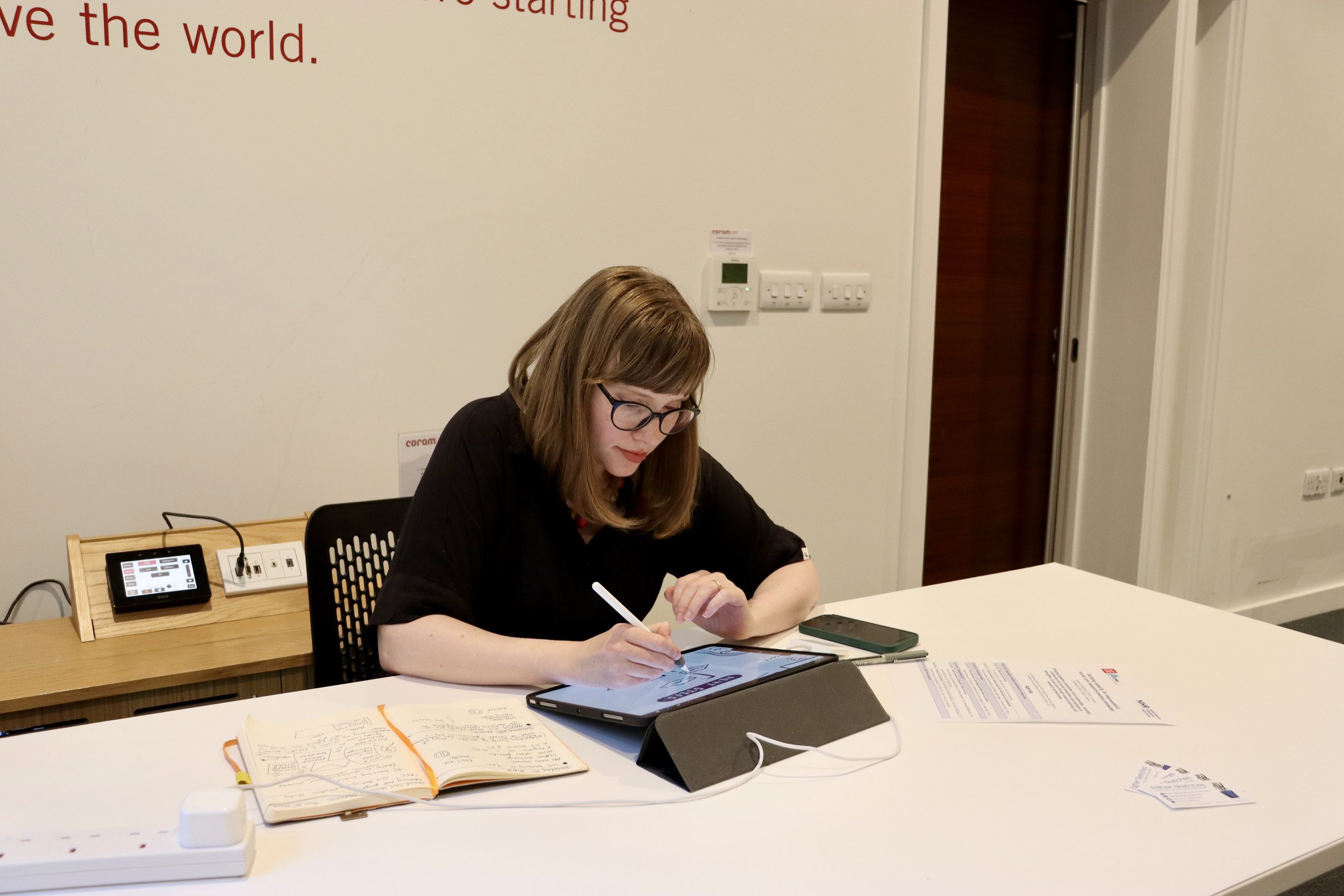 Esther is wearing a black top and sitting at a long desk working on her iPad. There is a notebook next to her on the desk.