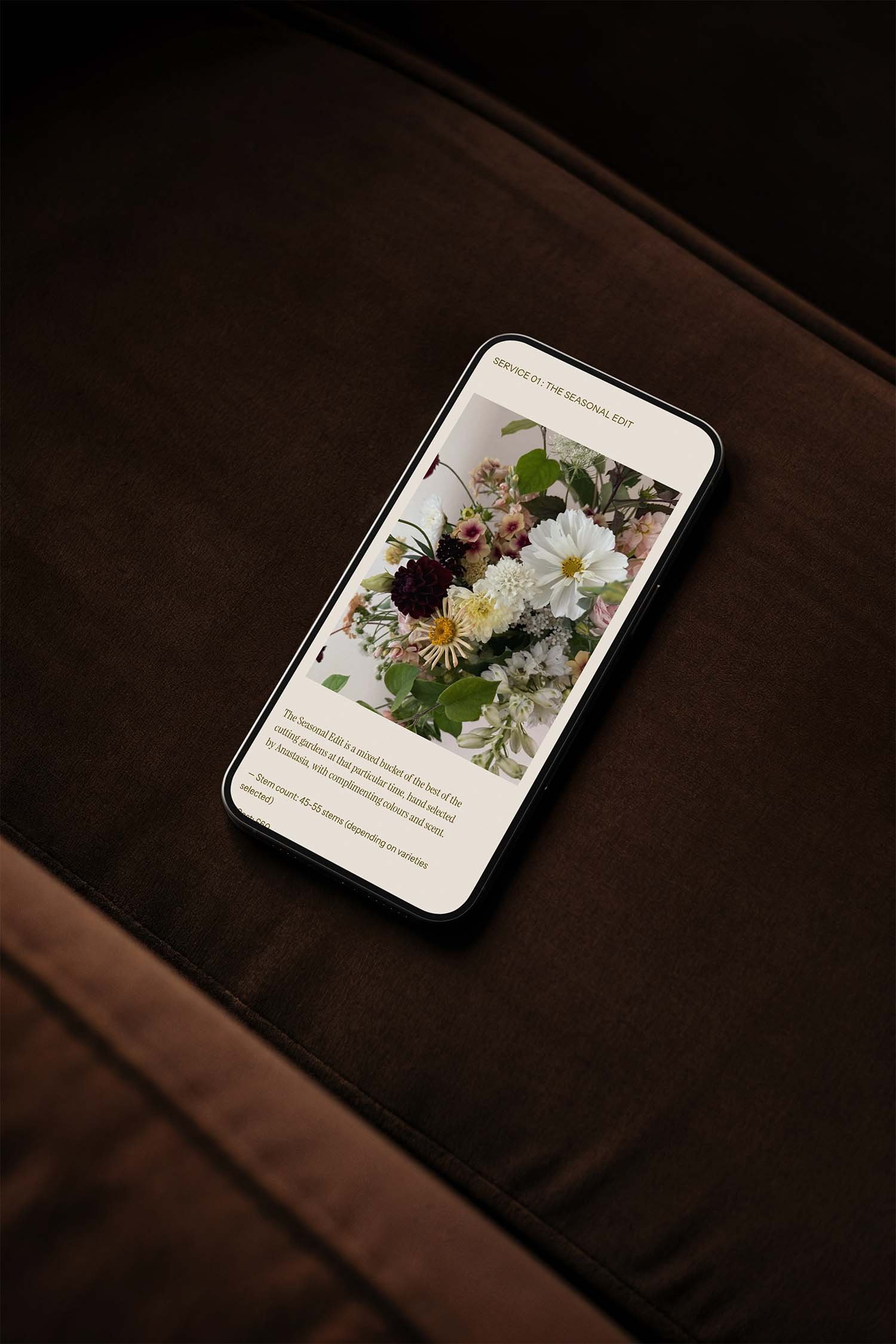 A smartphone on a brown upholstered surface displays an image of a flower arrangement with a description of a seasonal bouquet titled 'Service 01: The Seasonal Edit'.