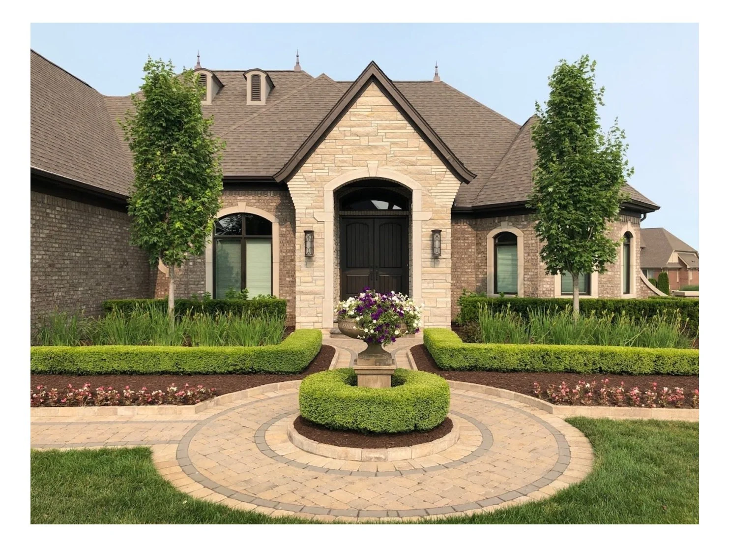 Front view of a large house with brick and stone exterior, landscaped garden, circular stone walkway, a fountain with flowers, and symmetrical trees.