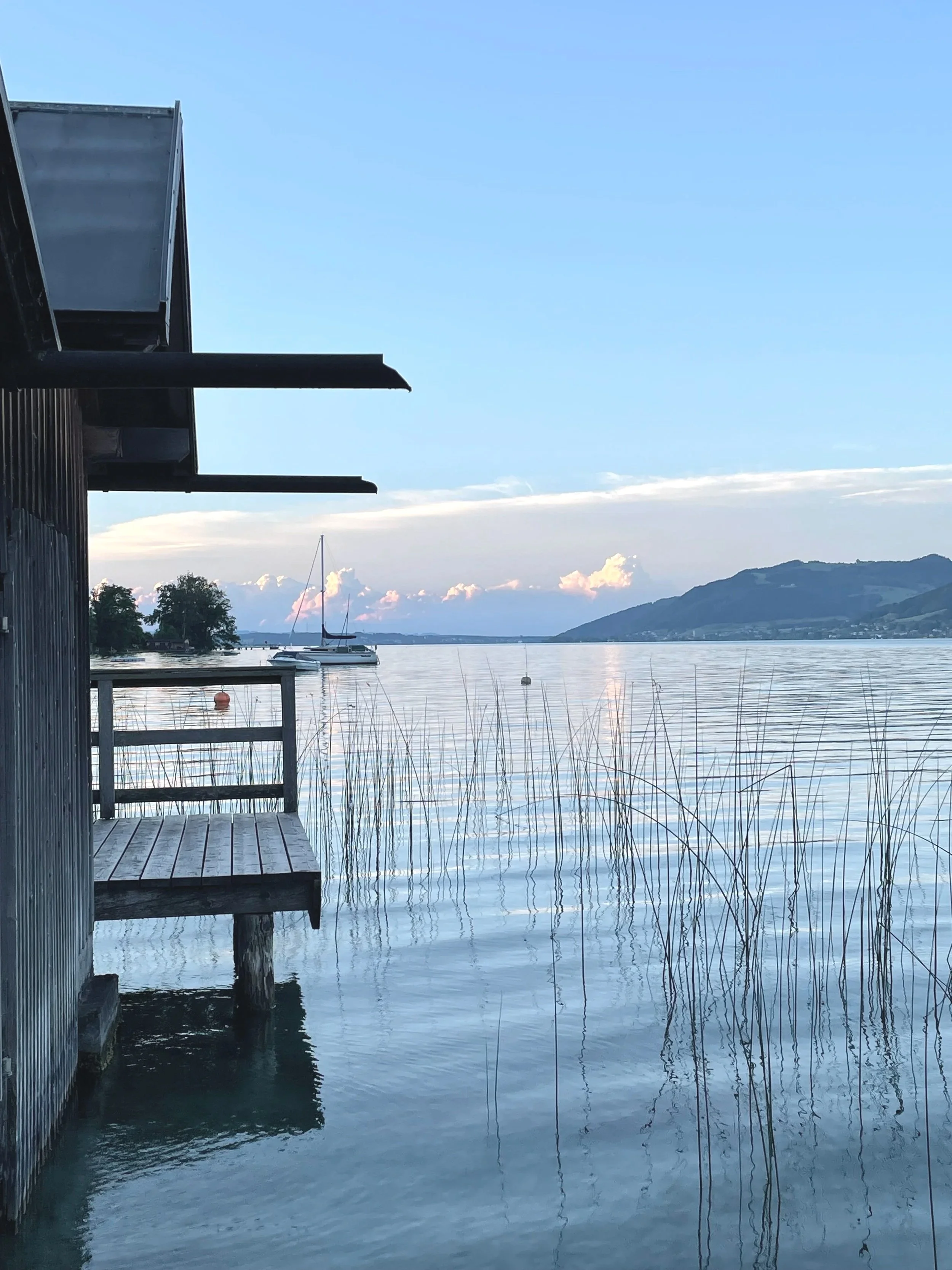 Blick auf einen See mit Booten, Berge im Hintergrund, ein Steg im Vordergrund, ein Gebäude mit Dach auf der linken Seite, die Weite des Wassers und die Wolken am Himmel.
