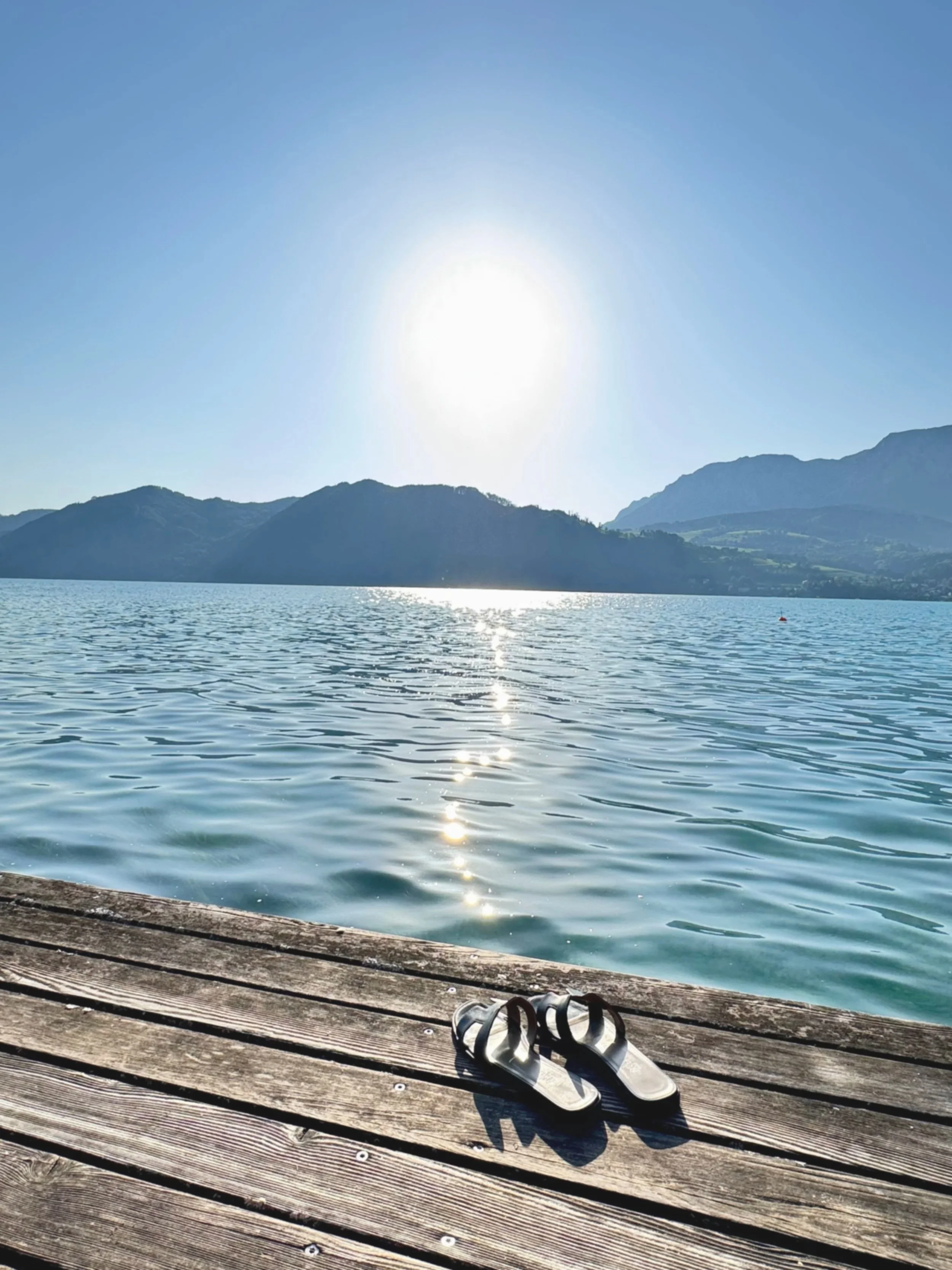 Blick auf einen See mit Bergen im Hintergrund, am Himmel die Sonne. Ein Paar Sandalen liegt auf einem Holzsteg am Ufer.