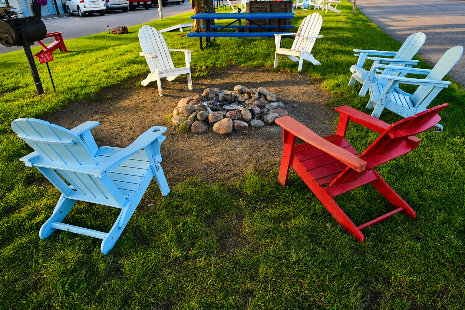 Outdoor campfire area at Vintage Block Inn & Suites in Okoboji, Iowa, featuring colorful Adirondack chairs, a stone fire pit, picnic table, and charcoal grill for guests to enjoy relaxing summer evenings.