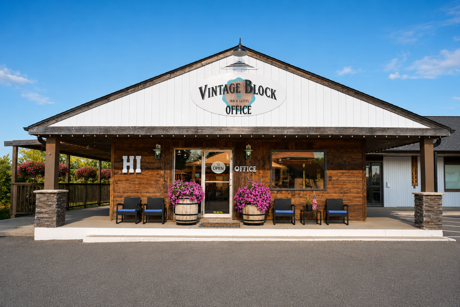 Front entrance of Vintage Block Inn & Suites in Okoboji, Iowa, featuring a rustic wood office with white siding, welcoming front porch, outdoor seating, colorful flower planters, and clear blue skies.