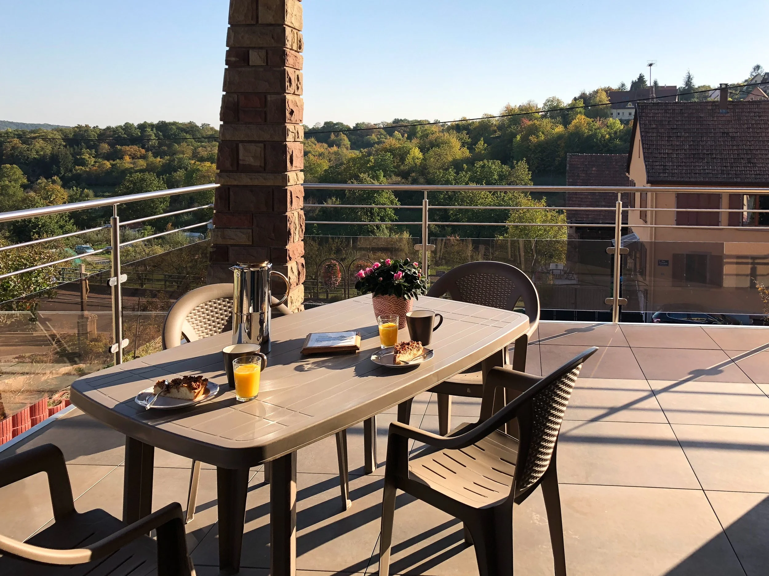 Table avec petits déjeuners et boissons, vue sur terrasse avec paysage vert et maisons, ciel clair, ambiance paisible.