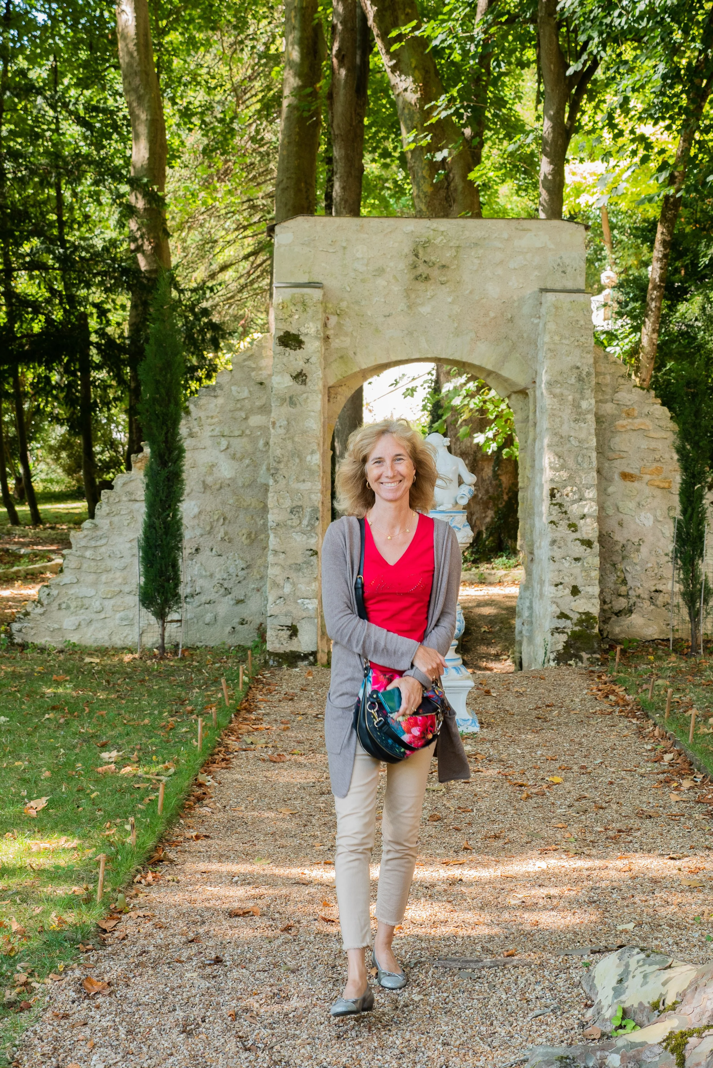 Une femme souriante portant un chemisier rouge, un gilet gris, un sac coloré et des chaussures à talons gris, marchant sur un chemin en gravier dans un parc avec une structure en pierre et des statues en arrière-plan.