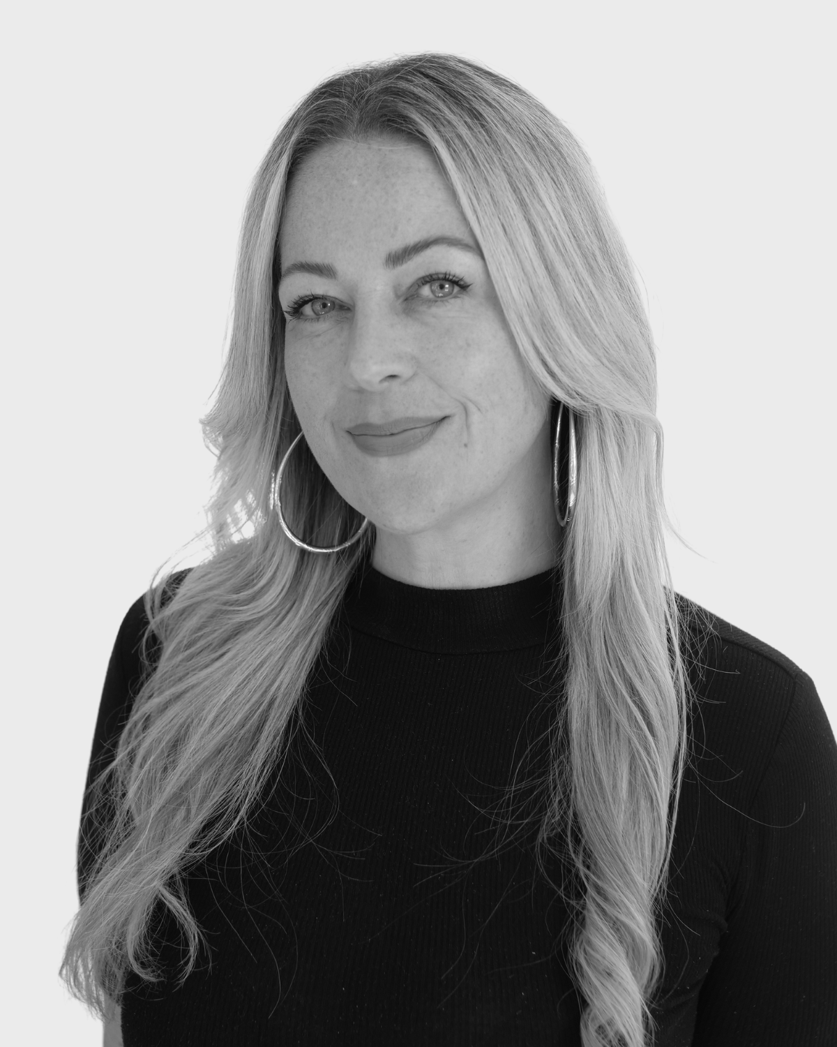 Black and white portrait of a woman with long, wavy hair, hoop earrings, and a confident smile, wearing a black top.