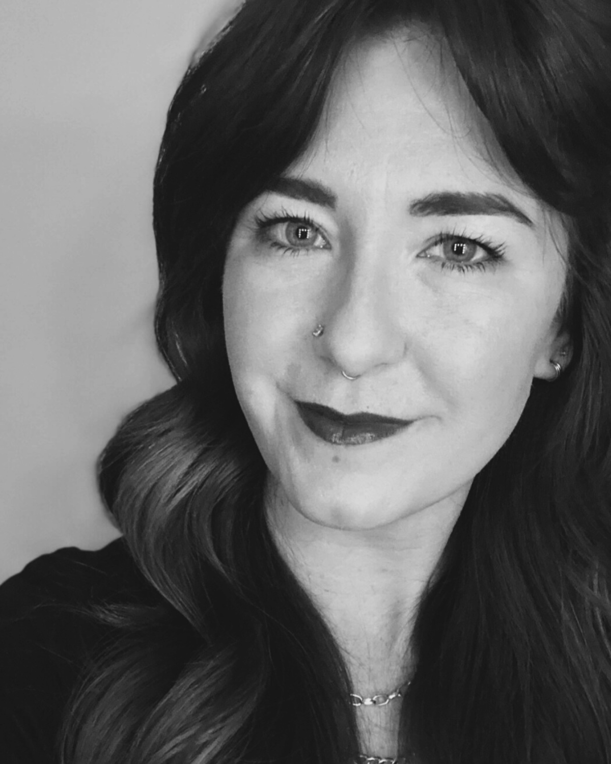 Black and white portrait of a young woman with wavy dark hair, light-colored eyes, multiple facial piercings, and wearing a chain necklace, smiling slightly.
