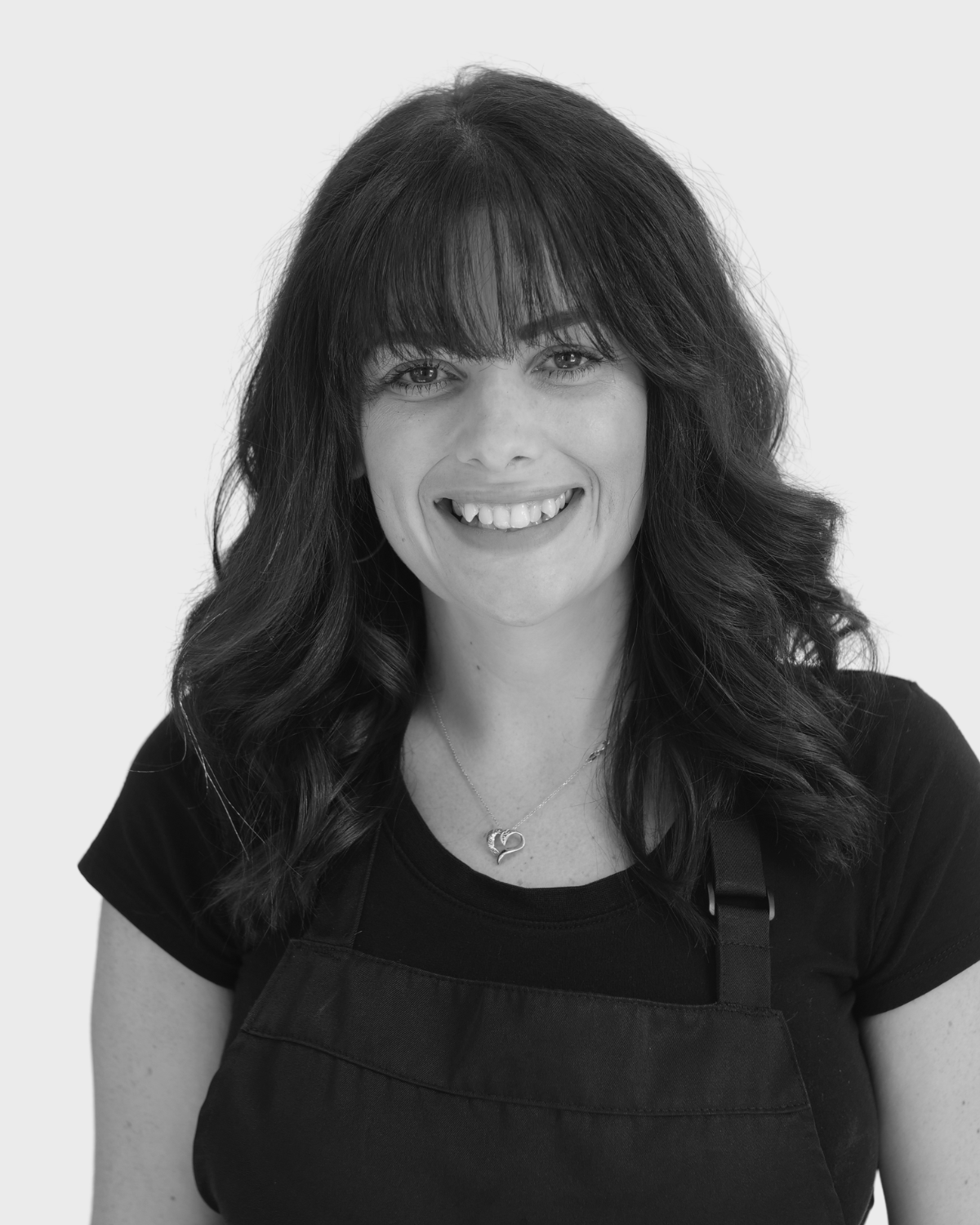 A woman with long, wavy hair and a fringe, wearing a black shirt and apron, smiling at the camera.