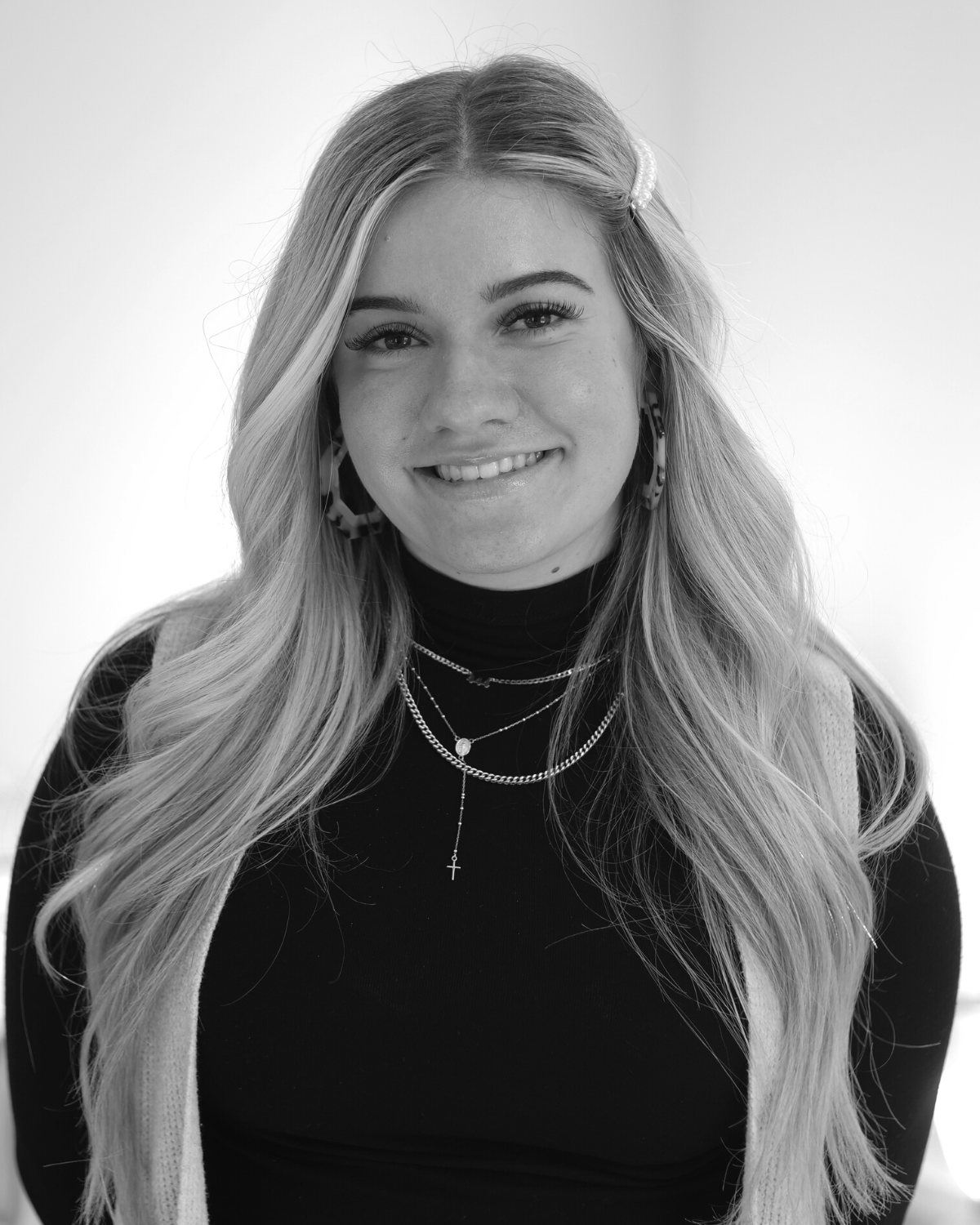 Black and white portrait of a young woman with long hair, wearing a turtleneck and layered necklaces, smiling at the camera.