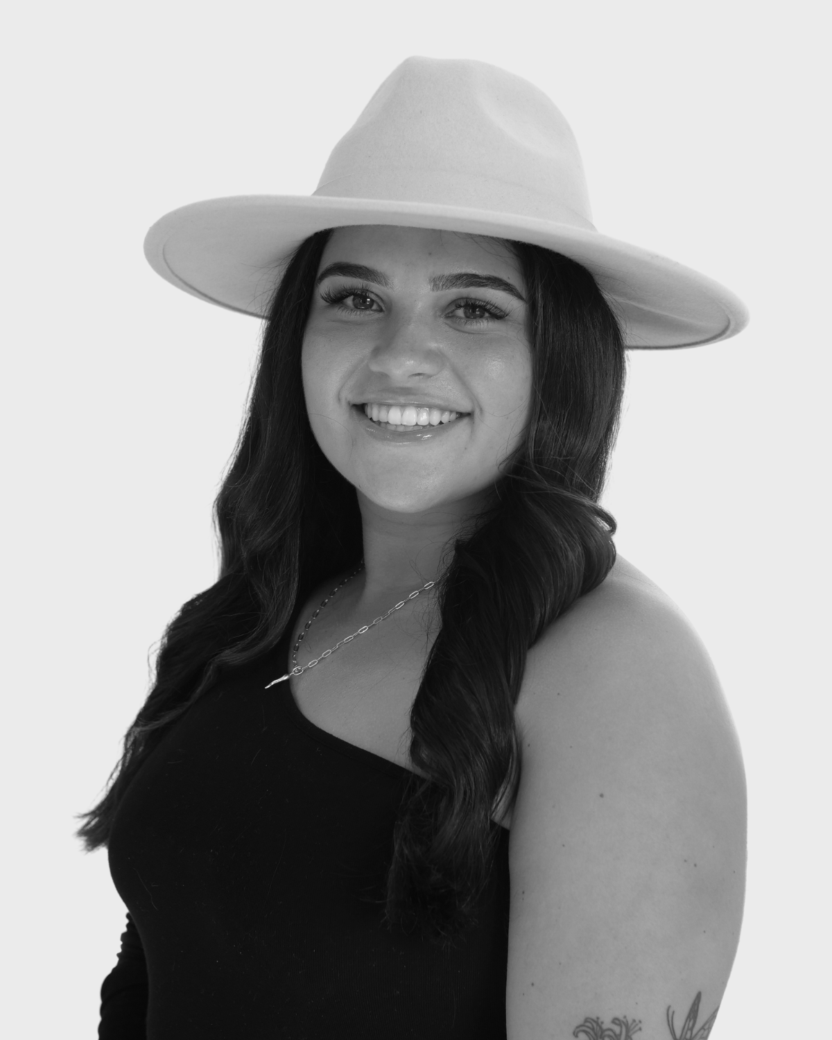 Black and white portrait of a smiling woman wearing a wide-brimmed hat and a black sleeveless top, with a tattoo on her arm and a necklace.