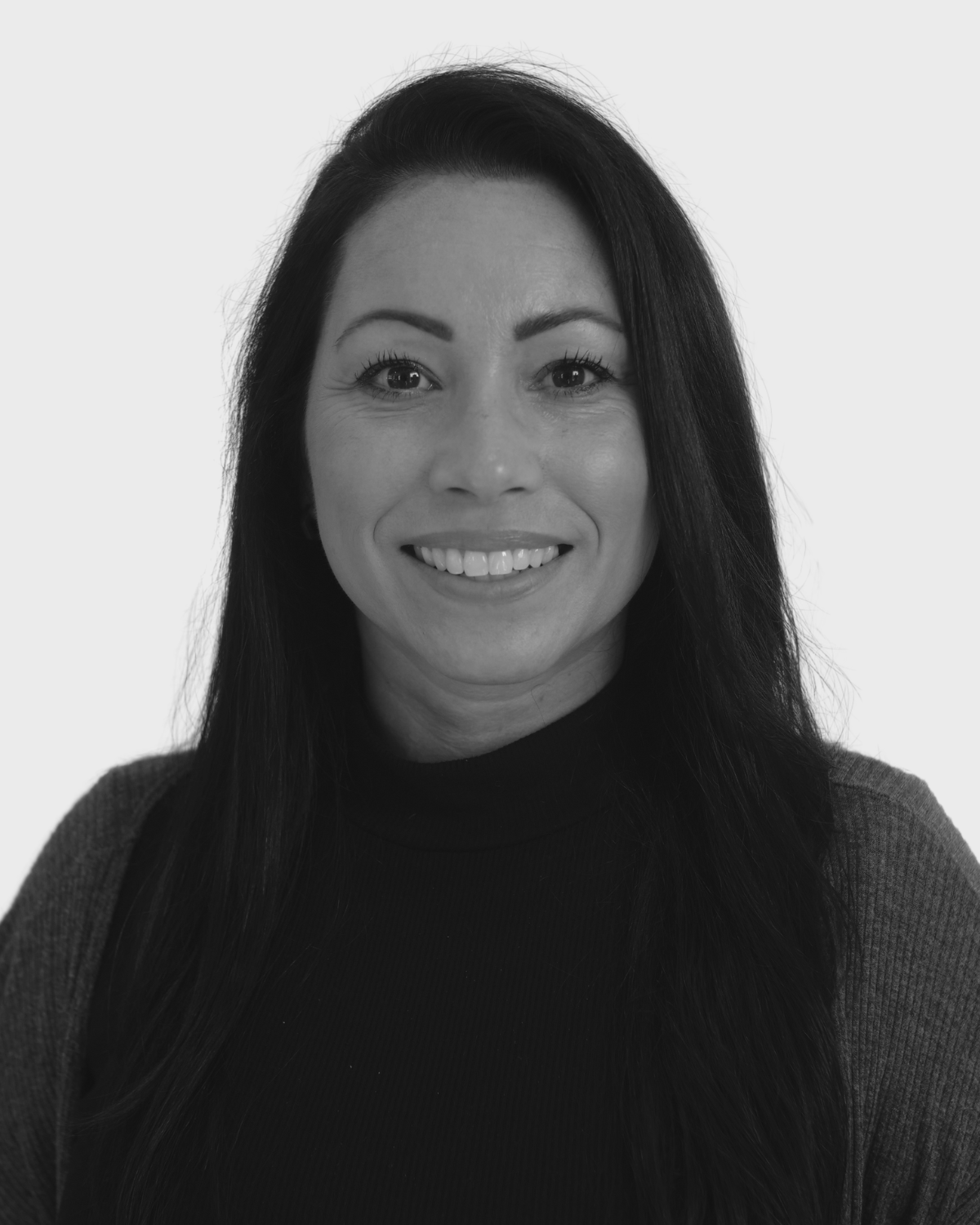 Black and white portrait of a smiling woman with long dark hair, wearing a dark turtleneck and a cardigan, against a plain light background.