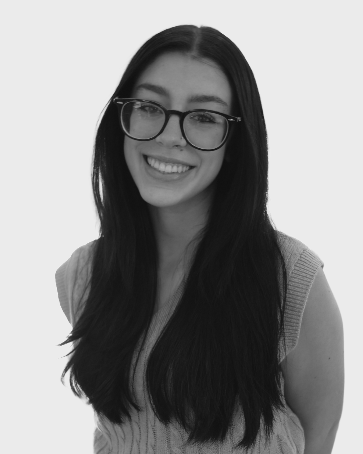 A young woman with long dark hair, large glasses, and a bright smile, wearing a textured sleeveless top against a plain background.