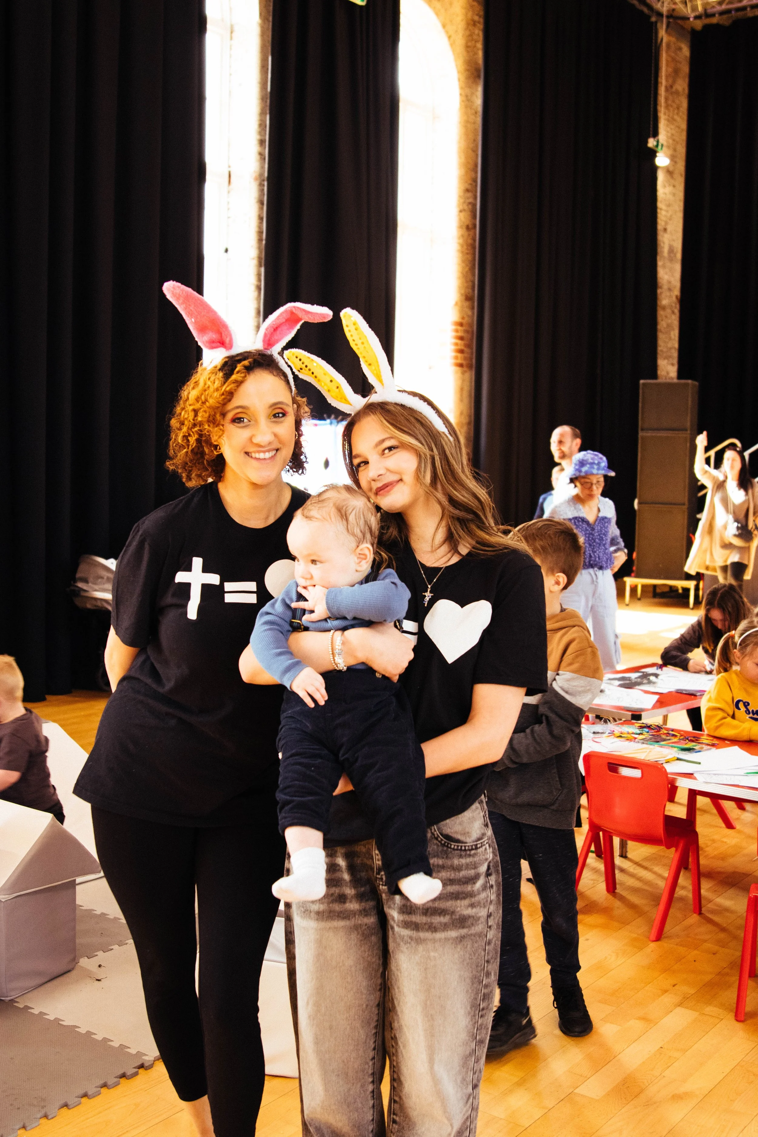 Two women wearing bunny ears hats smiling, with a baby in front of them, at an indoor event with children and tables in the background.