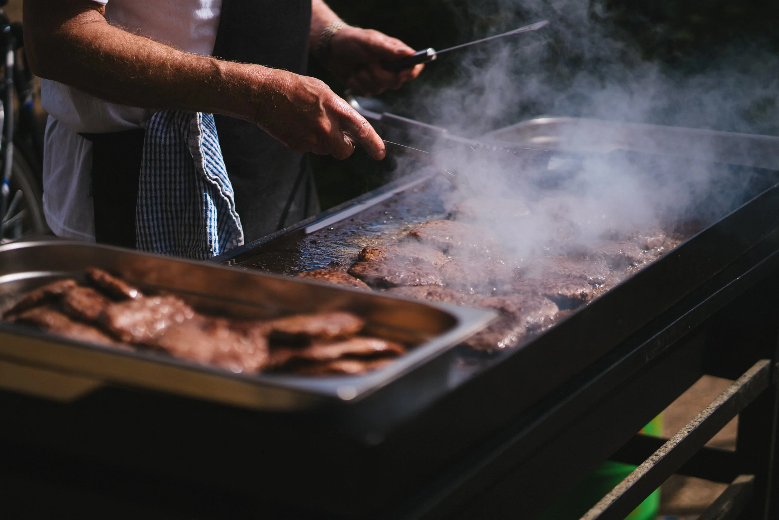 Two people cooking steaks on a large outdoor grill with smoke rising from the meat.