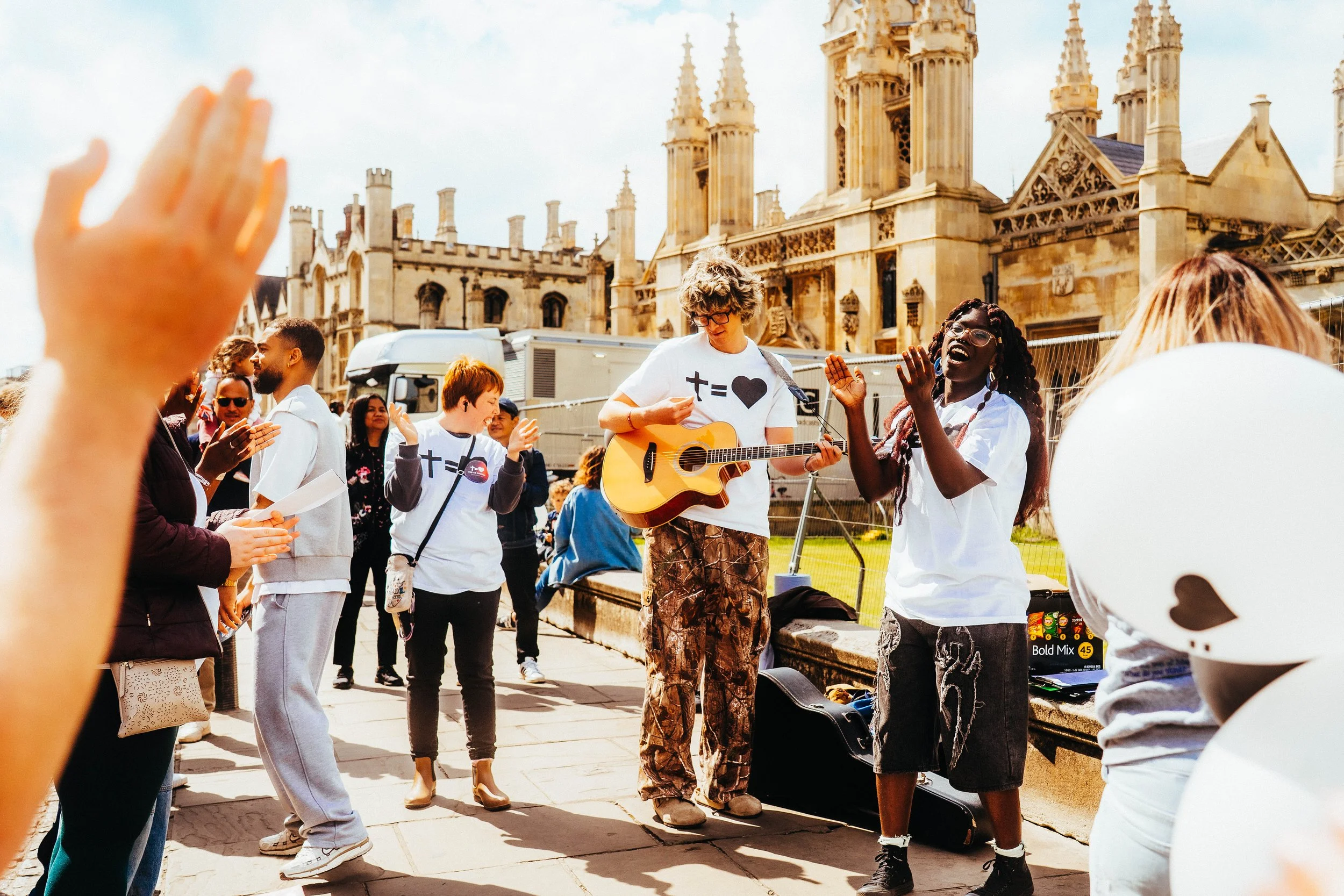 A group of people gathered outdoors near a historic building, with one person playing guitar and others singing or clapping, engaging in a lively street performance.