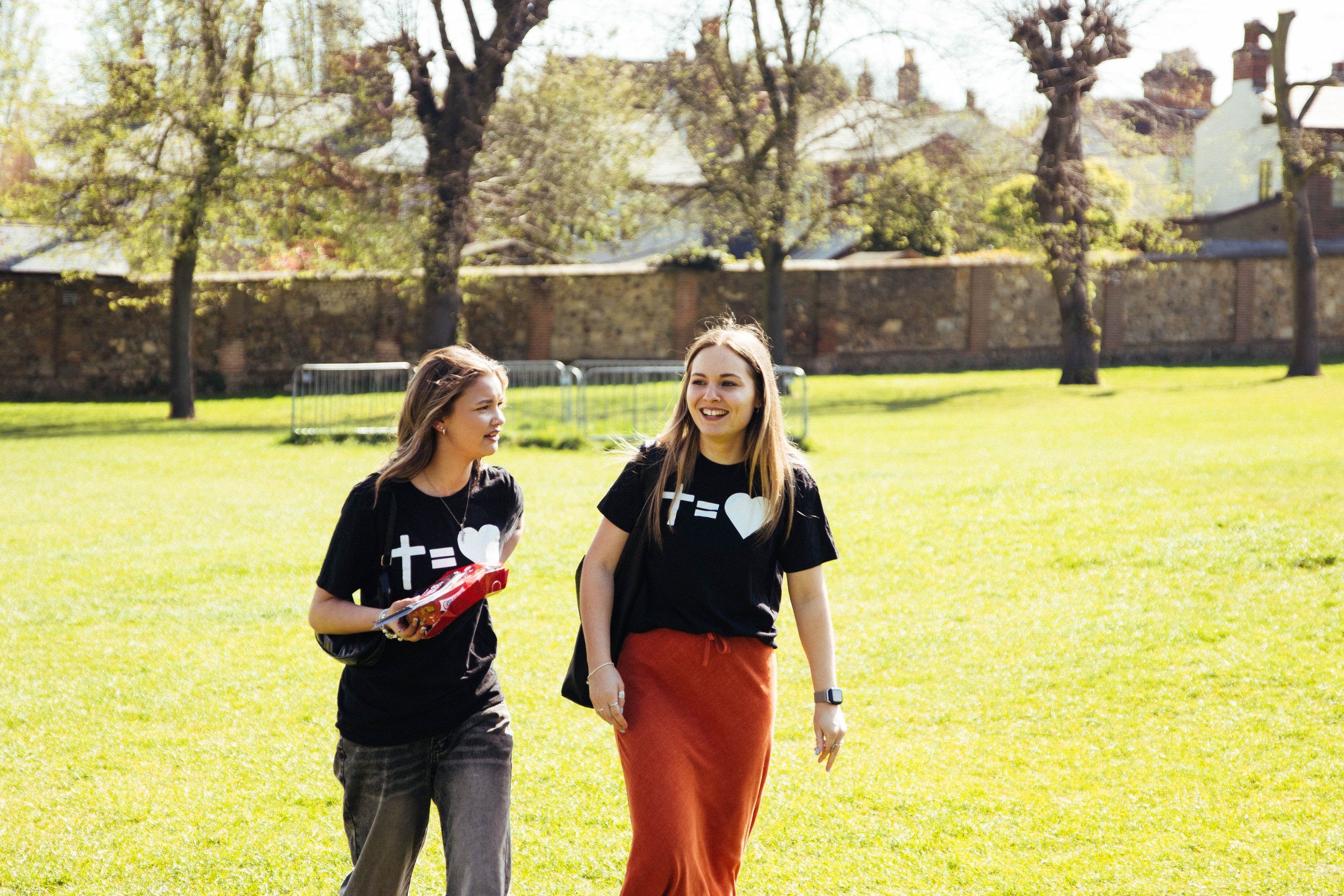 Two young women walking and smiling in a grassy park on a sunny day, wearing matching black T-shirts with hearts and equations.