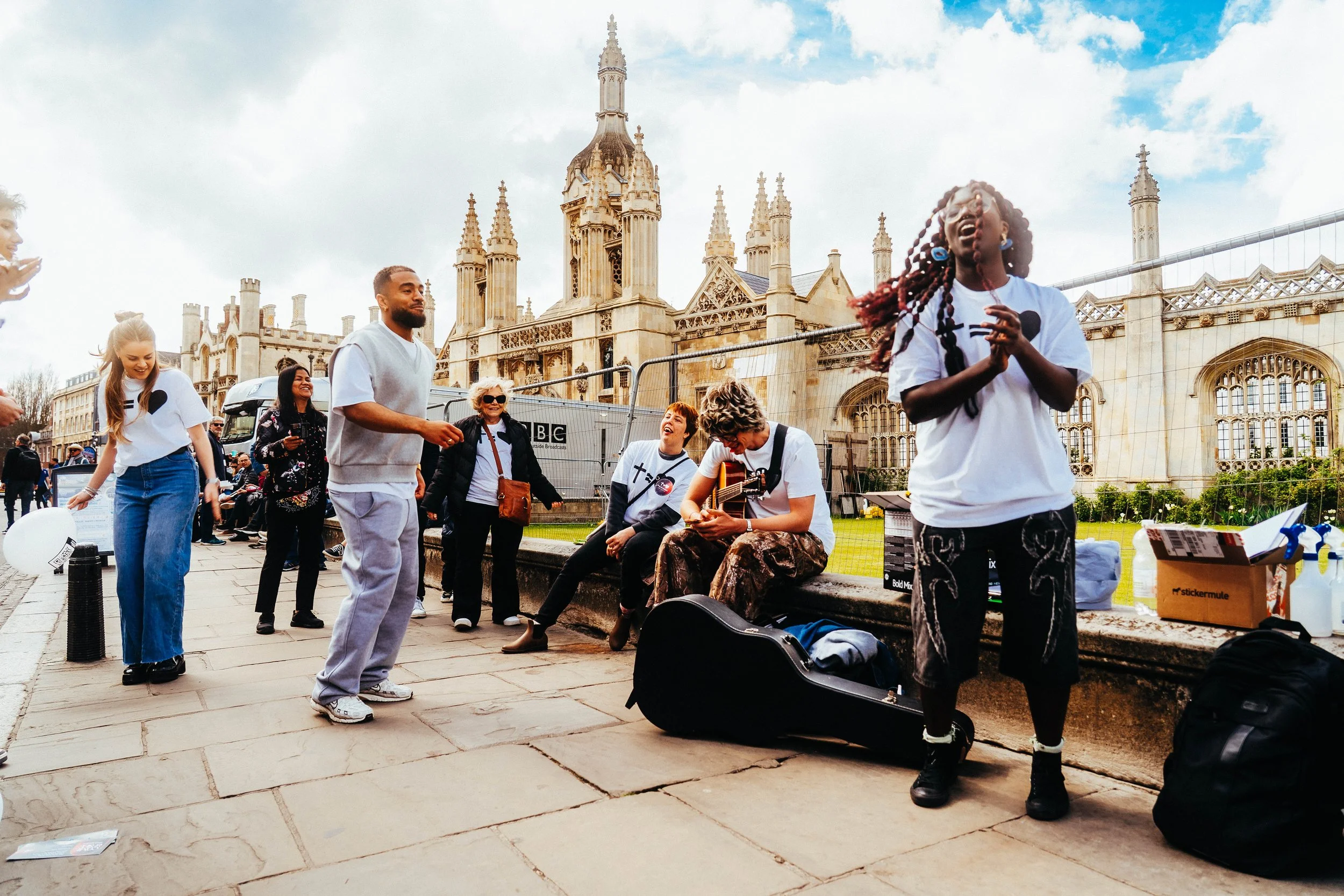 People gathered around a street performer singing and playing guitar outside of a historic building with gothic architecture, in a city square on a partly cloudy day.