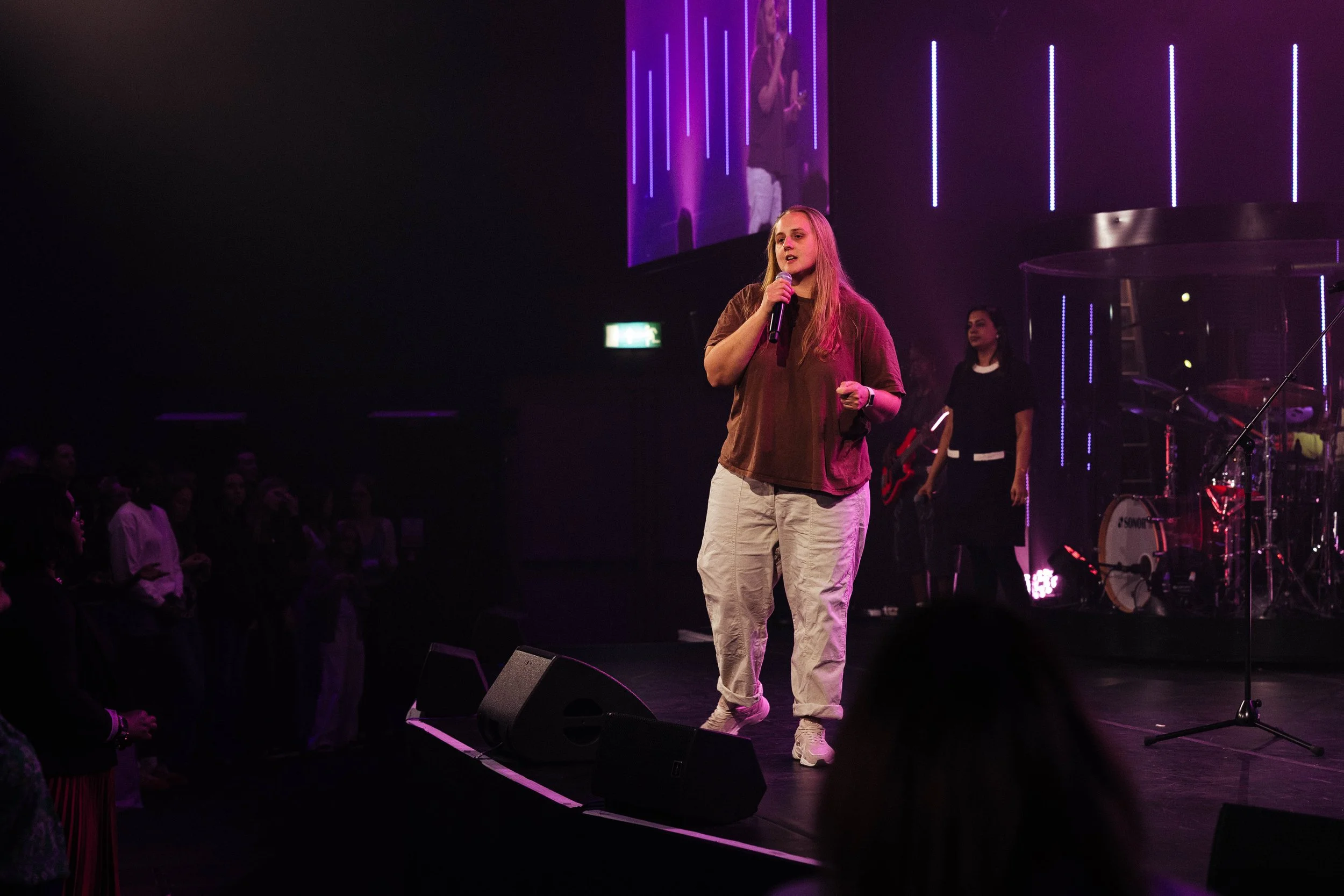 A woman with long blonde hair holding a microphone on stage, speaking to an audience in a dark room with purple and pink lights, behind her are musicians with instruments and a large screen displaying her image.