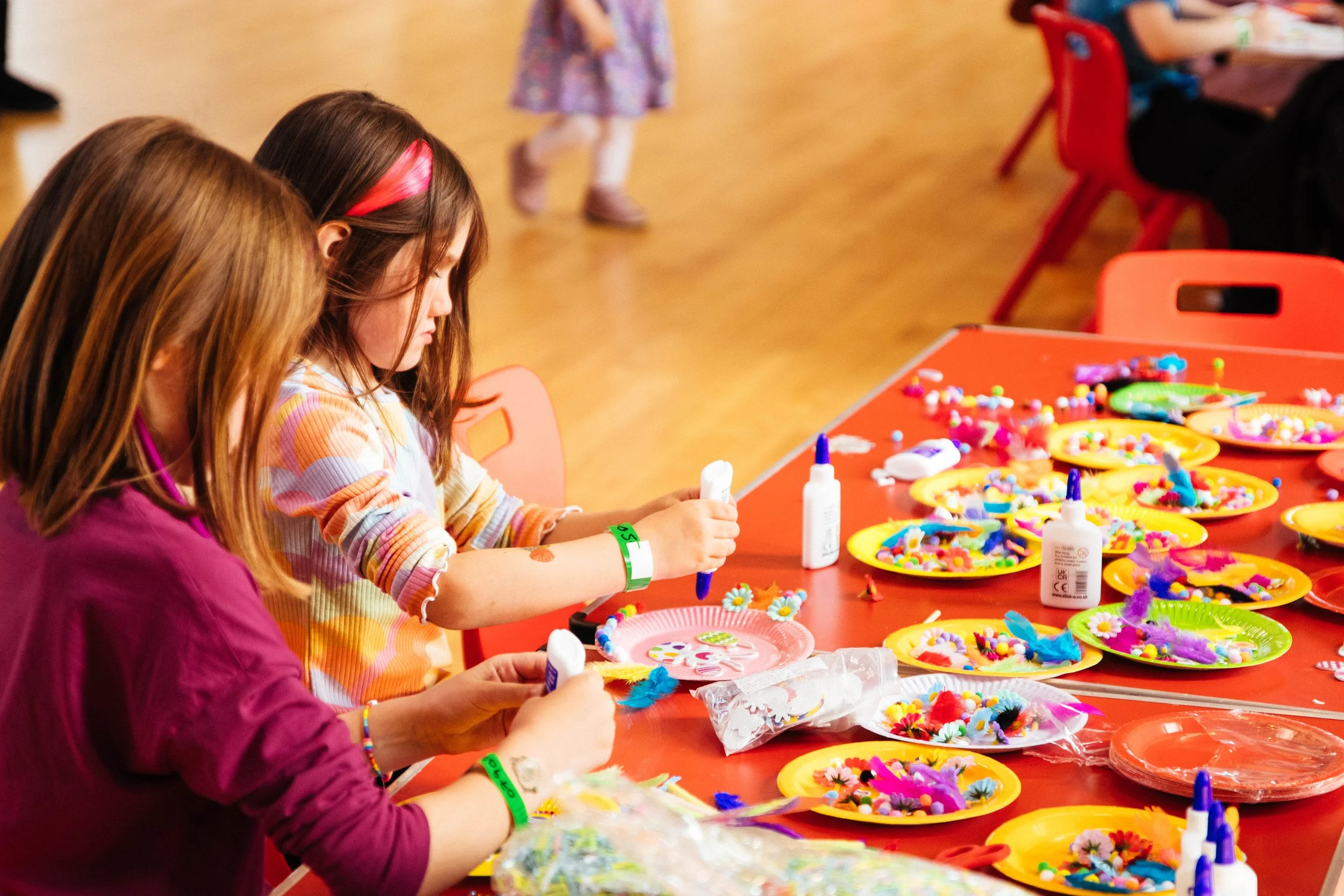 Children creating holiday crafts at a table with colorful decorations, feathers, and glue.
