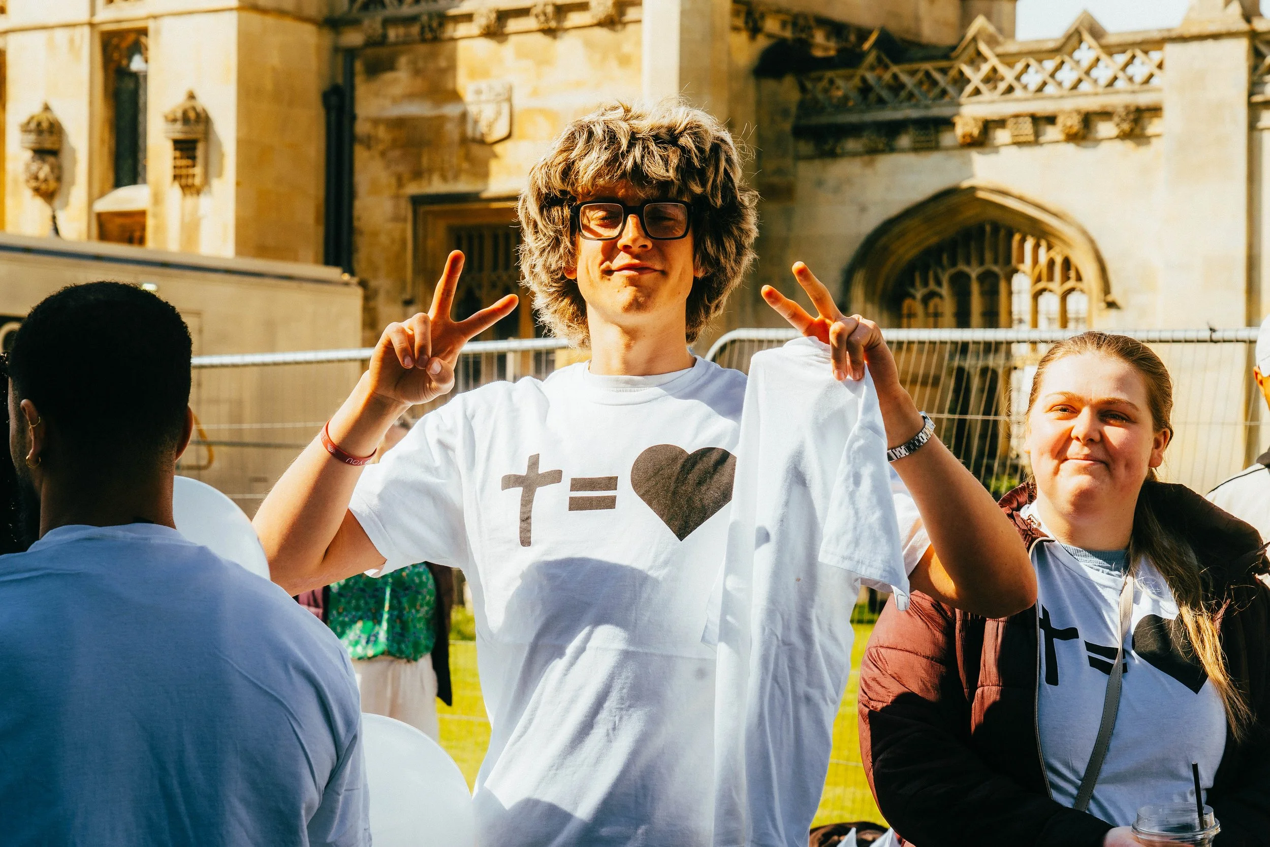 A young man with curly hair, glasses, and a white t-shirt displaying 't = heart', making peace signs with both hands; a young woman next to him wearing a similar shirt and holding a drink, outdoors in front of an old stone building.