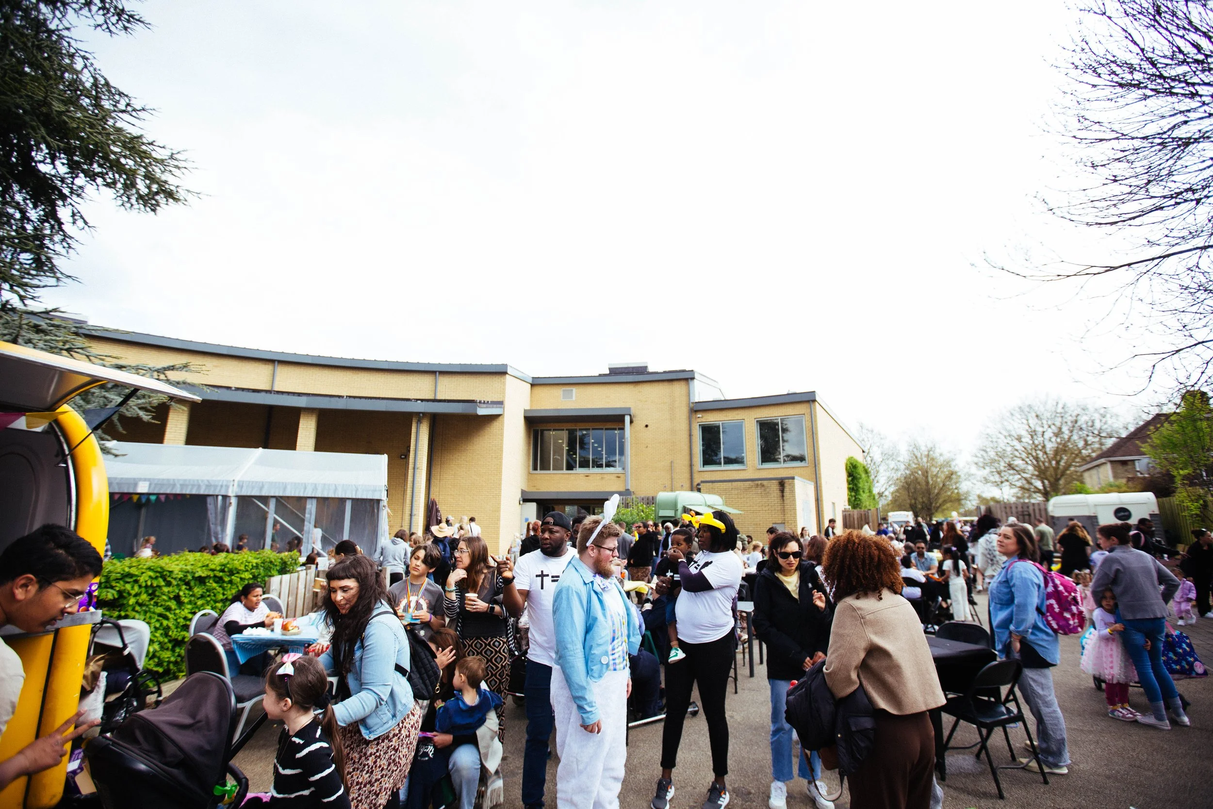 Crowd of people at an outdoor event or gathering in a residential area, with children and adults engaging in activities, and a large yellow and black inflatable structure on the left side of the image.